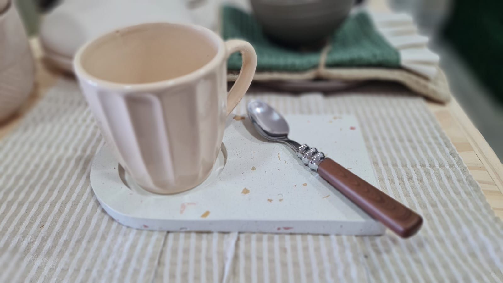 Taza beige sobre una bandeja blanca con una cuchara sobre un mantel a rayas.