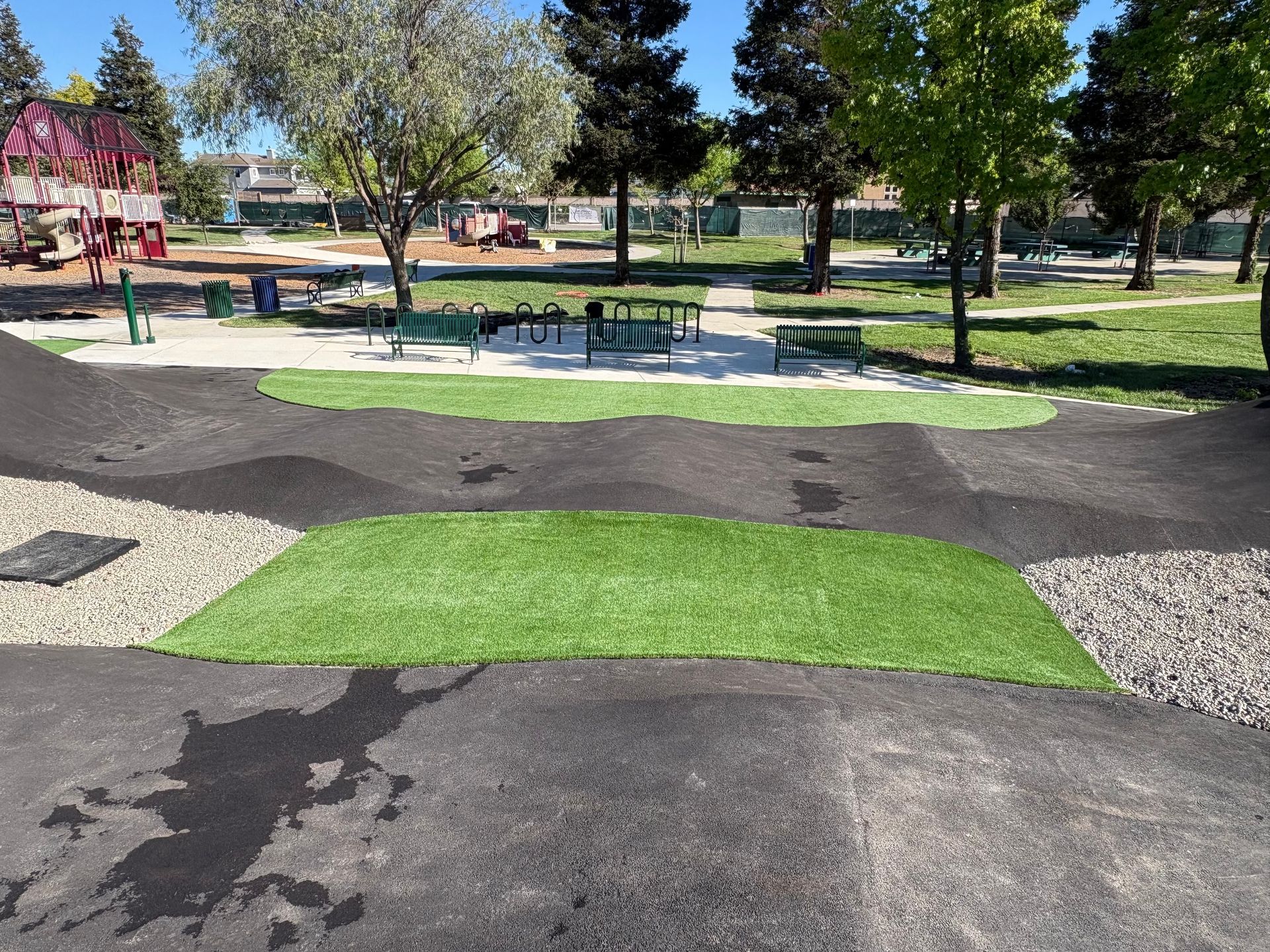 Park with artificial turf paths, trees, and picnic tables in bright daylight.
