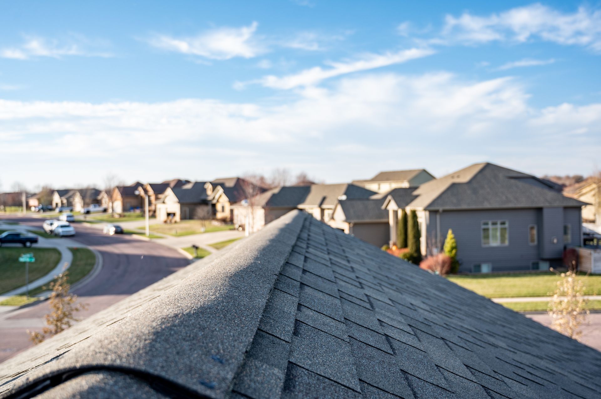 Residential shingle roof ridge overlooking suburban neighborhood under blue sky.