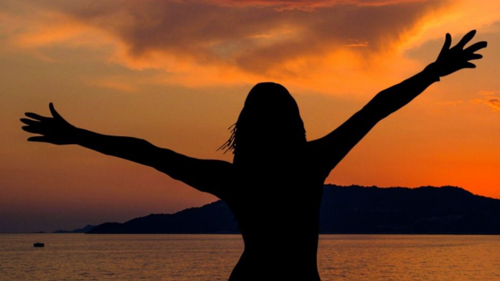 Silhouette of a person with arms raised, facing a sunset over water.