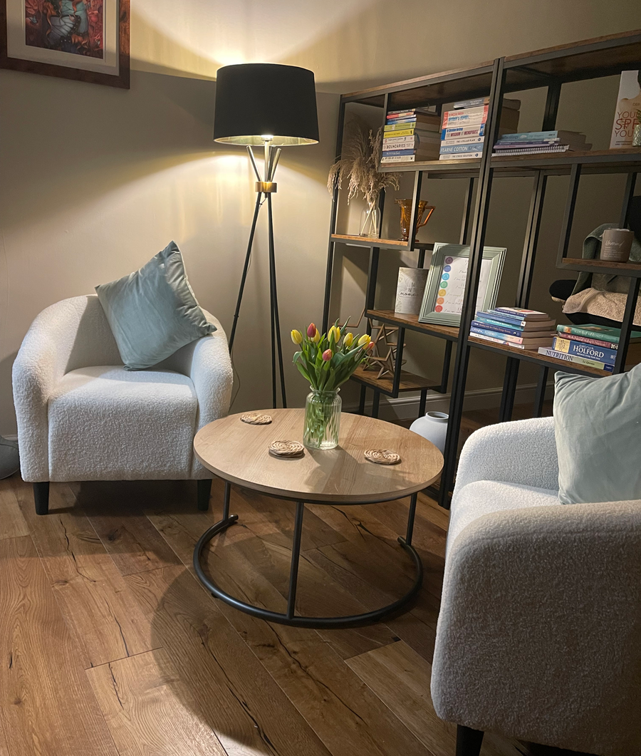 Cosy seating area with two fluffy armchairs, a round wooden table, a floor lamp, and a bookshelf. At Sage Therapies in Heaton