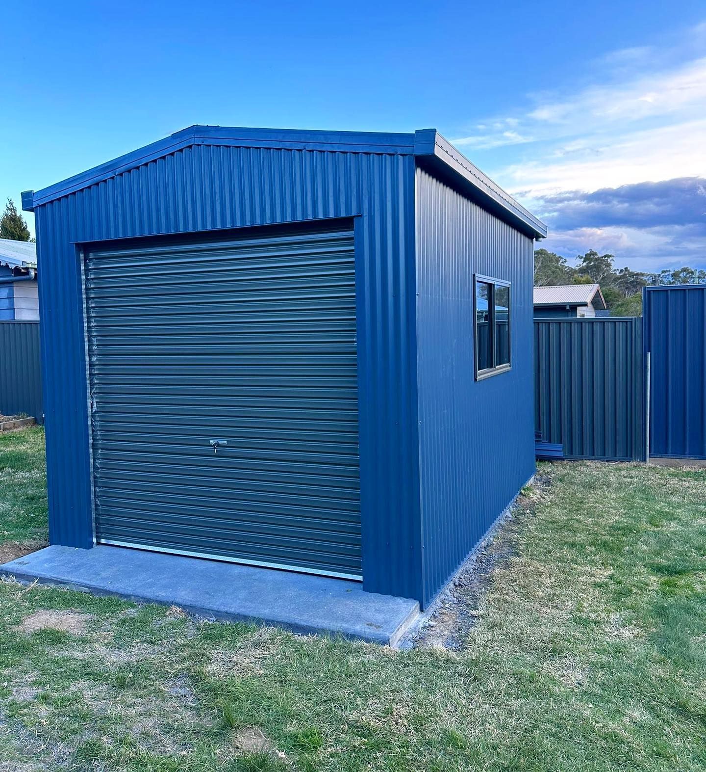 Blue Corrugated Metal Shed with A Grey Roller Door and Small Window, on A Concrete Slab — Complete Sheds In Marulan, NSW