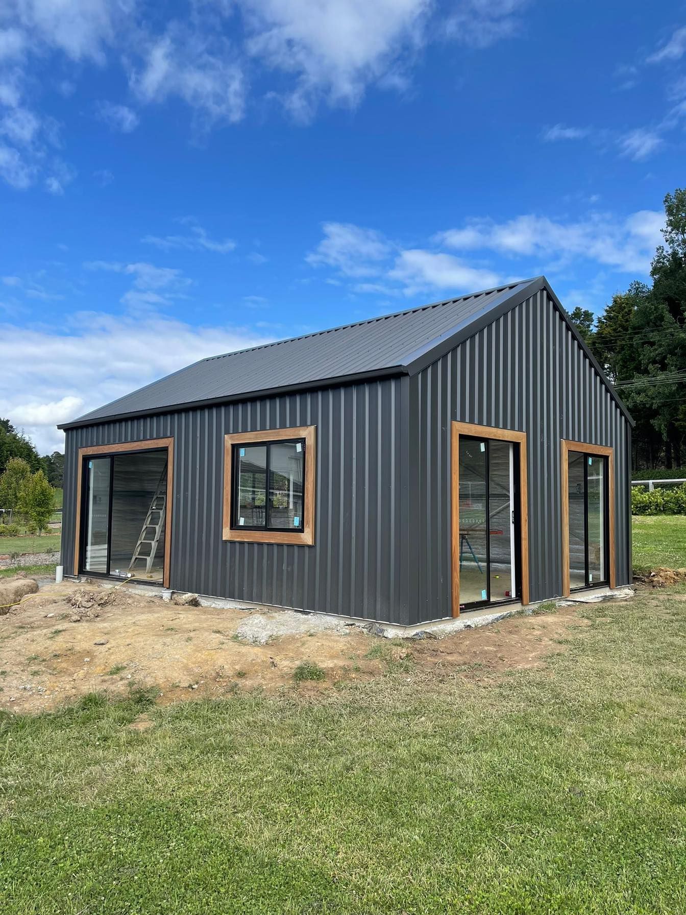 Dark grey corrugated metal building with wooden window frames, set on green grass under a blue sky.