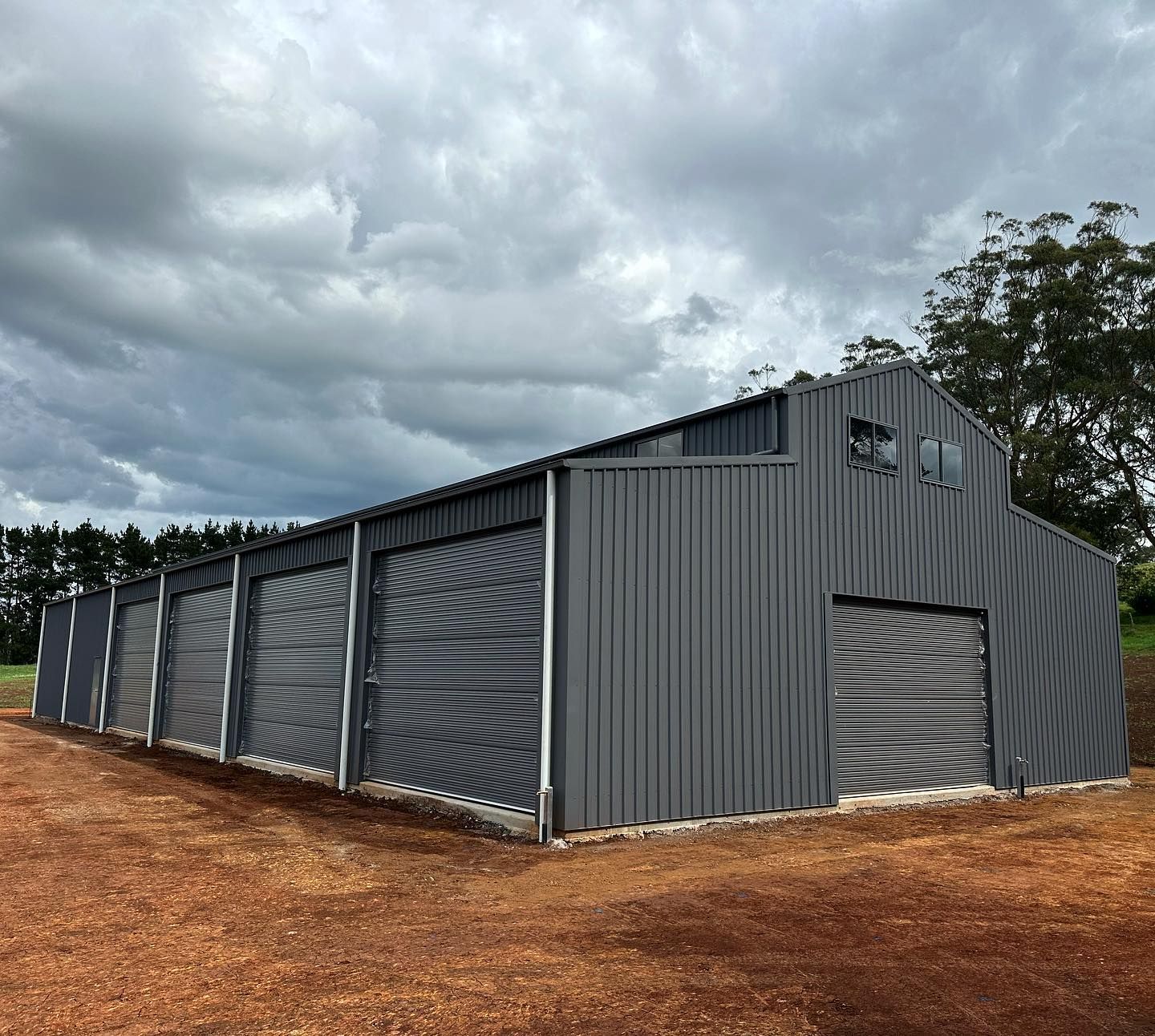 Grey Metal Building with Multiple Garage Doors Under a Cloudy Sky — Complete Sheds In Marulan, NSW