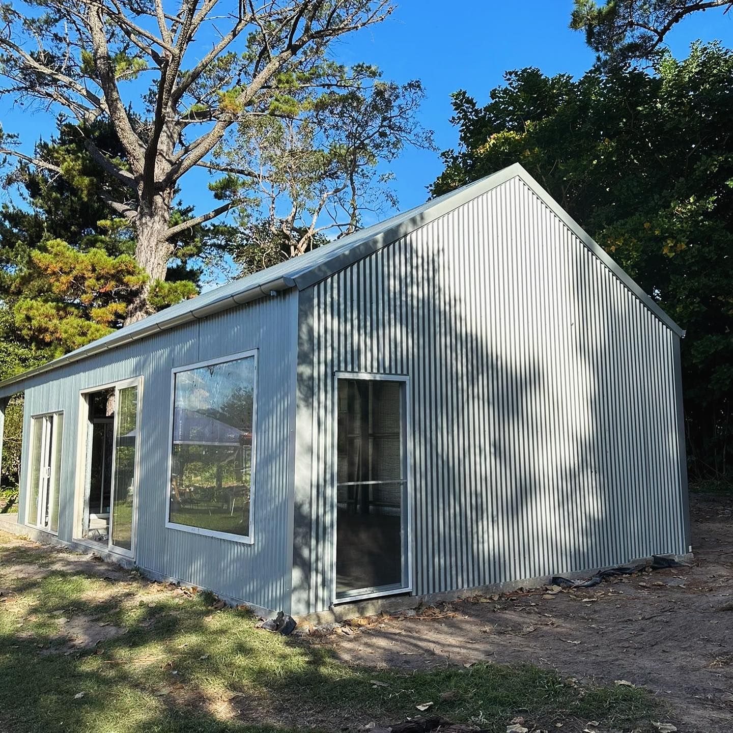Grey corrugated metal shed with large windows, set amidst trees under a blue sky.