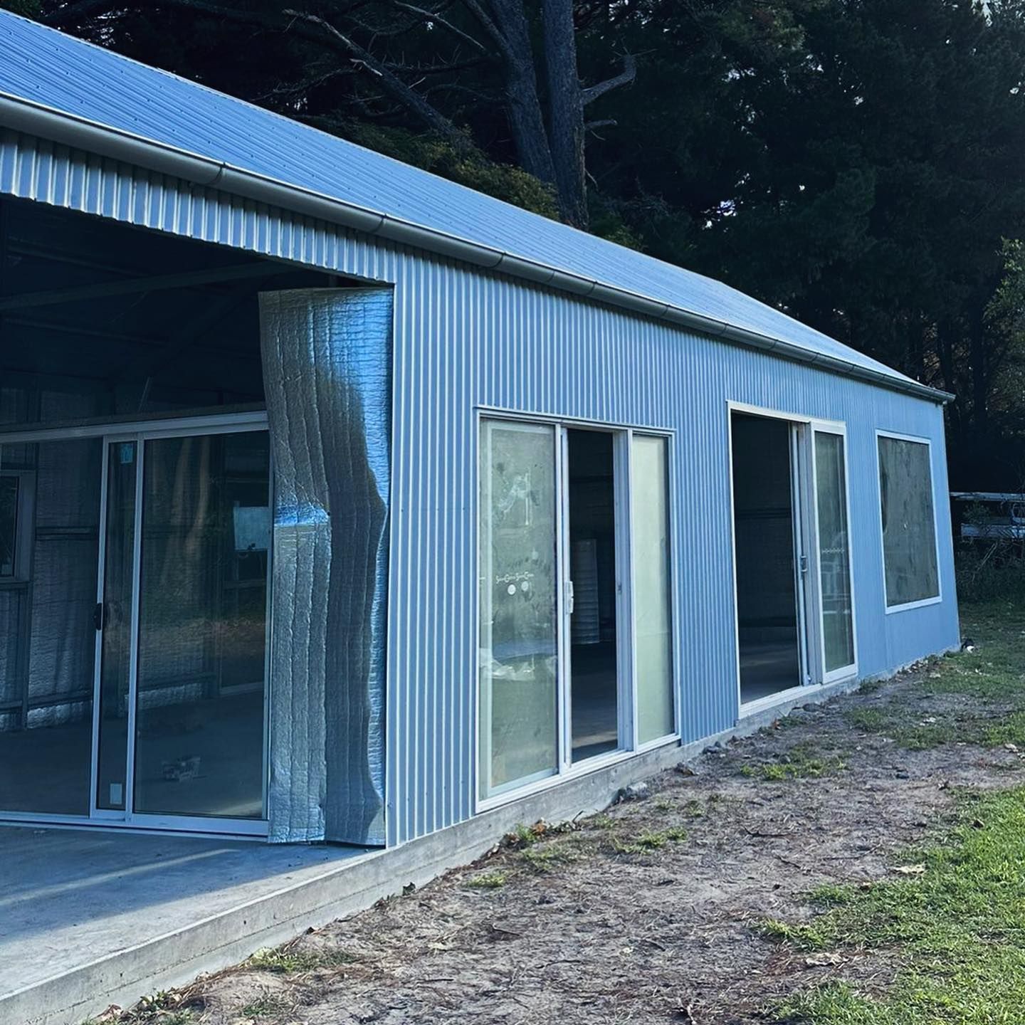 Exterior of a corrugated metal building with glass sliding doors and windows on a concrete slab, outdoors.