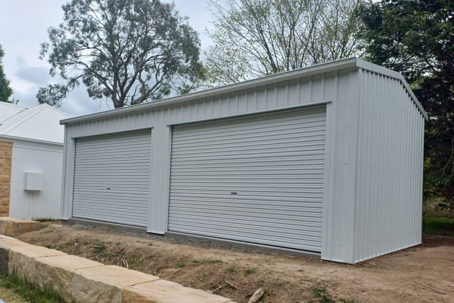 White metal two-car garage with roll-up doors, set on a gravel base beside a small building, outdoors.