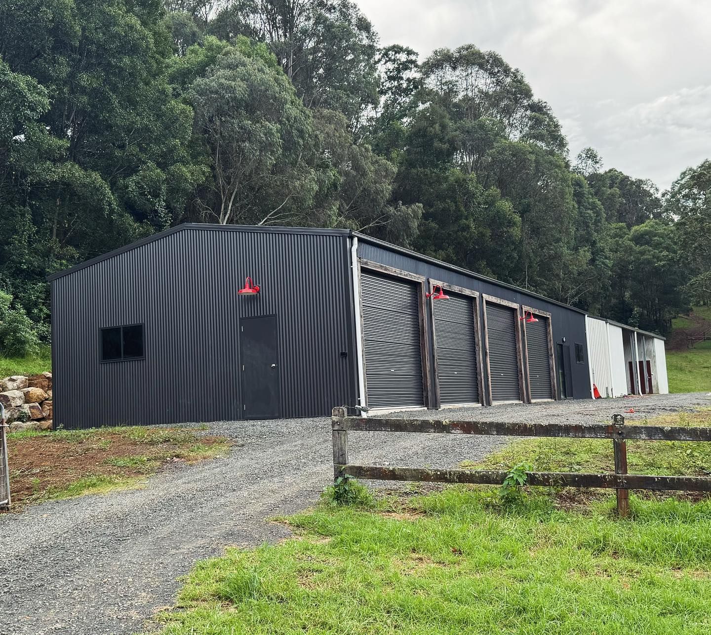 Black corrugated metal building with several garage doors, set in a grassy area with trees.