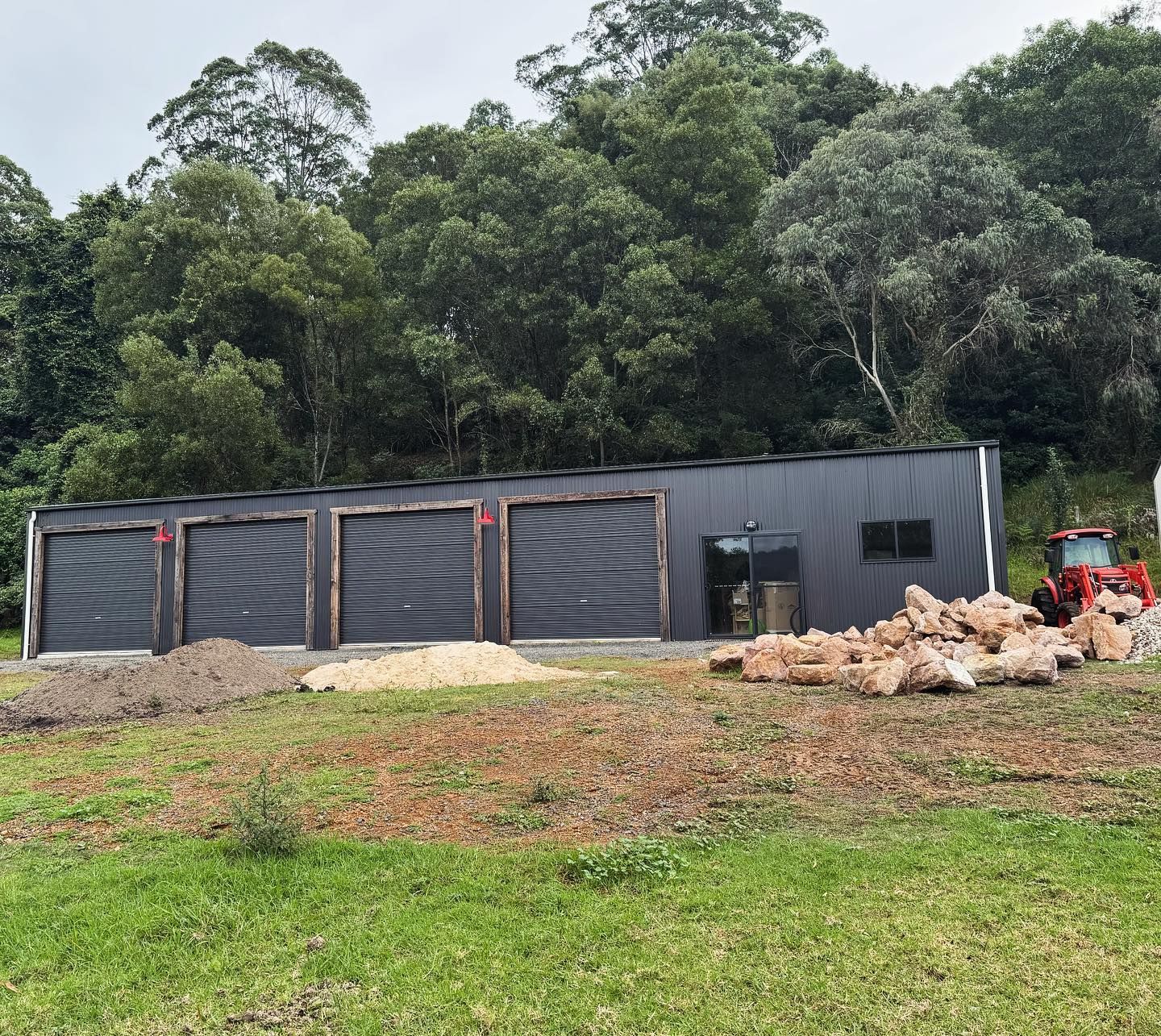 Grey garage with three bays and wooden accents; small tractor parked nearby. Green hillside in background.