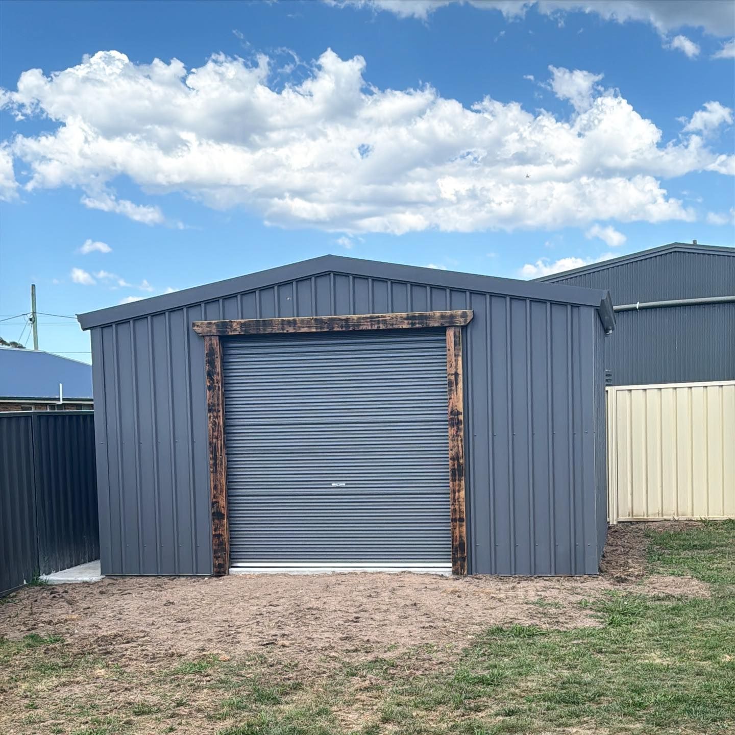 Grey shed with a roll-up door, framed with wood, in a yard under a cloudy blue sky.