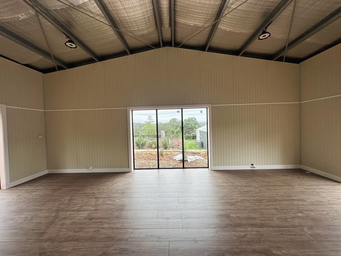 Interior of a large, empty room with light wood flooring, white walls, and a large glass door leading outside.