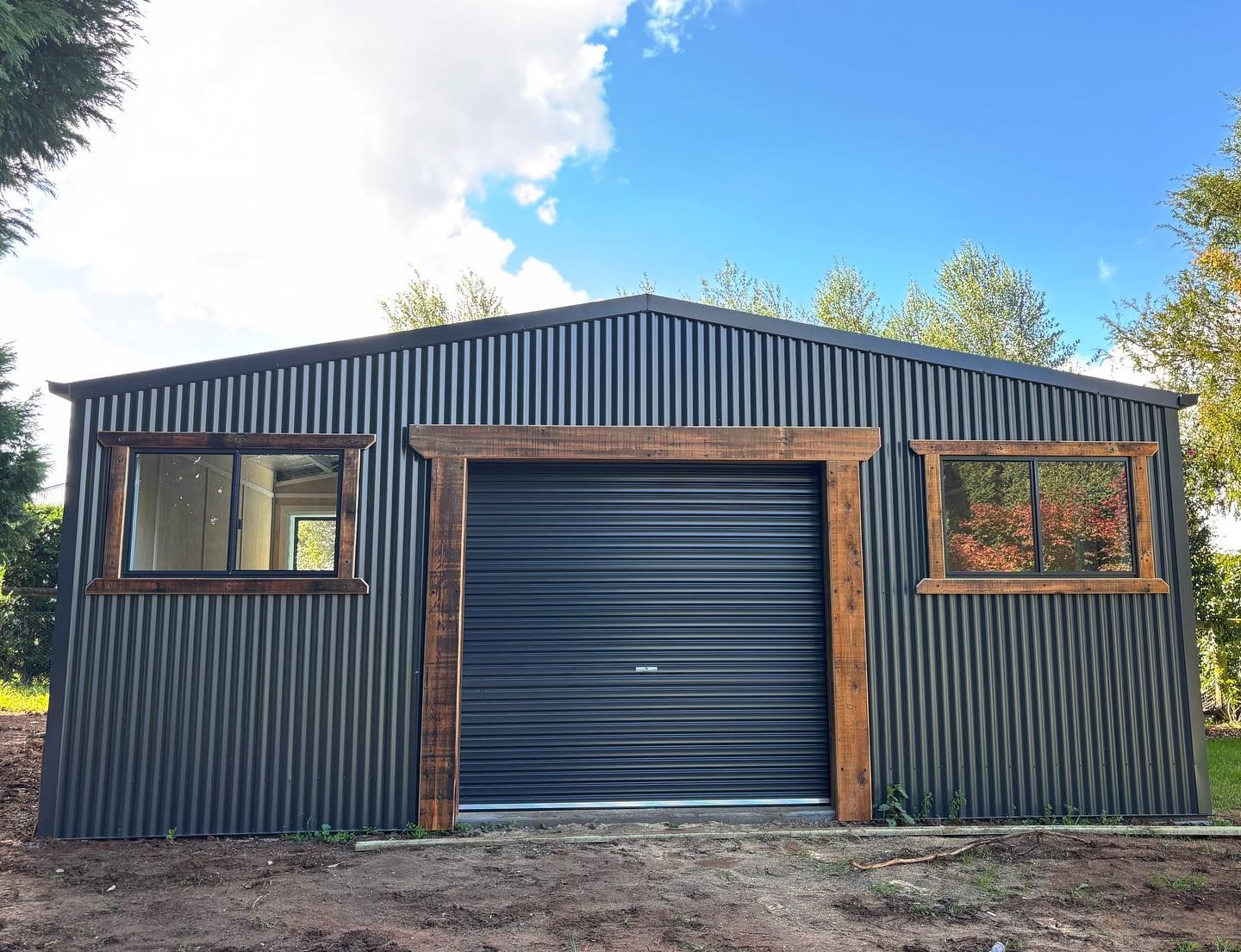 Dark grey corrugated metal shed with black garage door, wooden window frames, and blue sky.