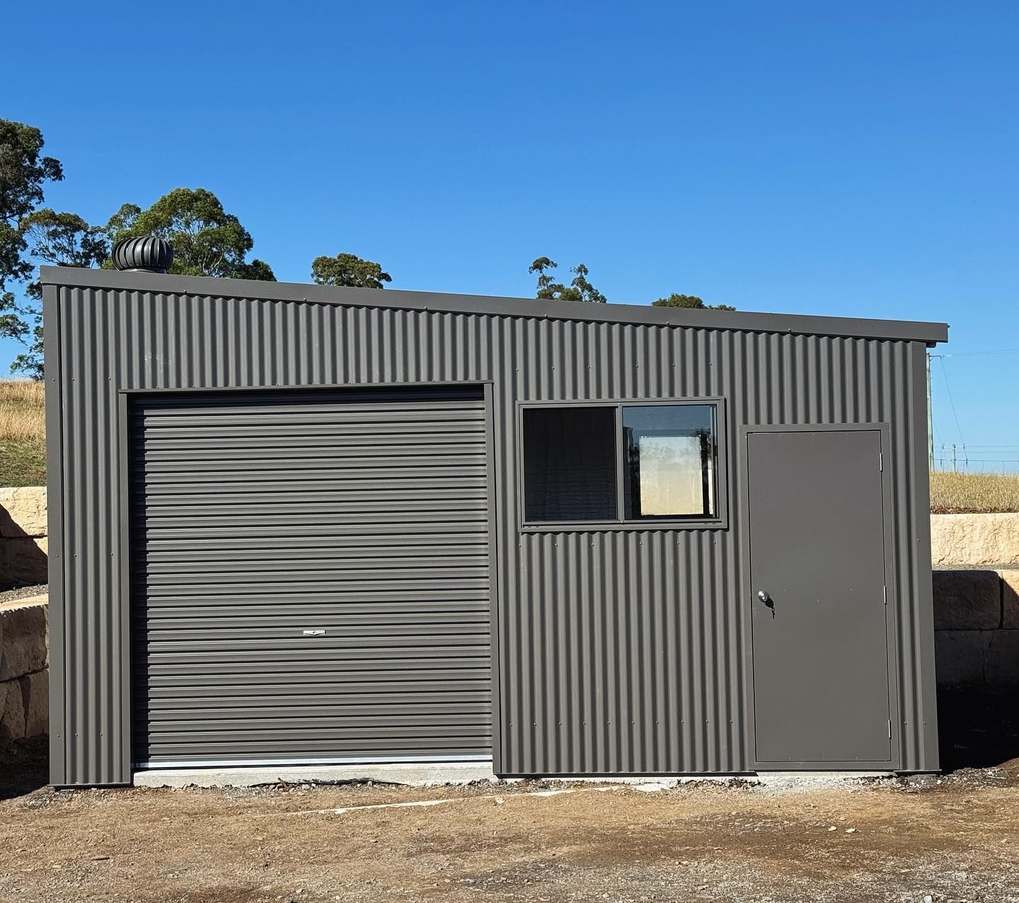 Grey corrugated metal shed with a roller door, window, and door against a blue sky.