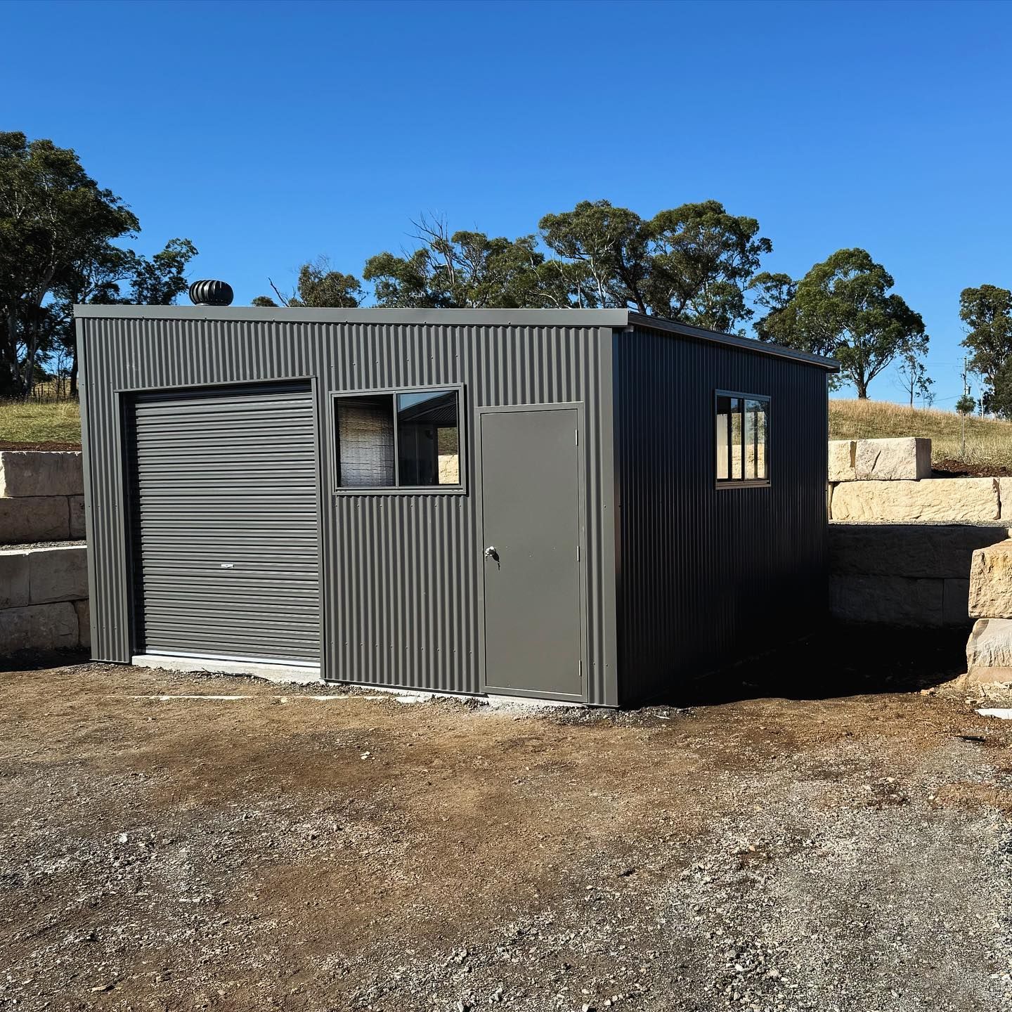 Grey corrugated metal shed with a roller door, window, and door, on a gravel surface under a blue sky.