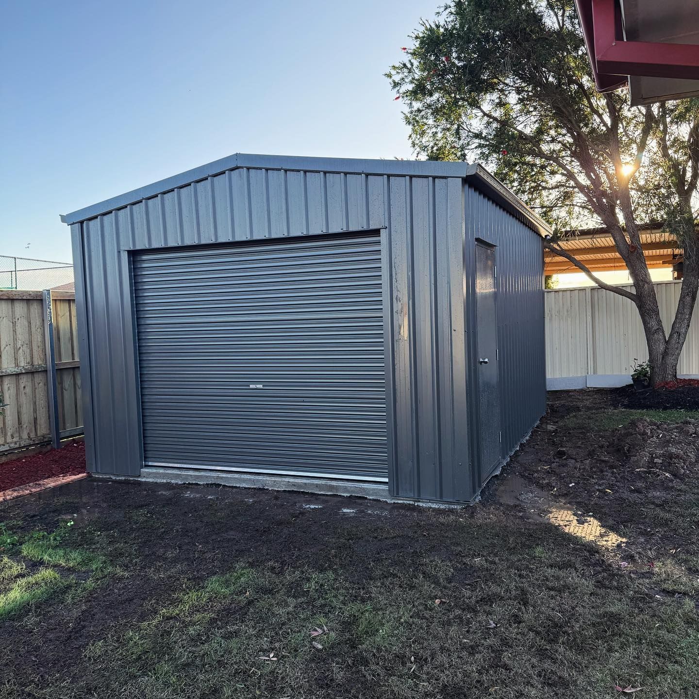 A grey metal shed with a roller door and side door, set in a yard.