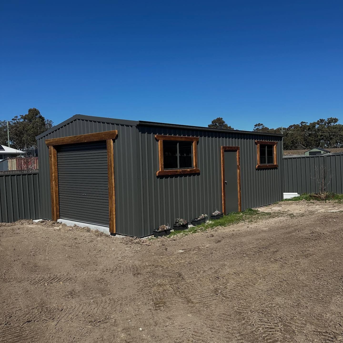 Grey metal shed with a roller door, windows, and a door, set in a gravel area on a sunny day.