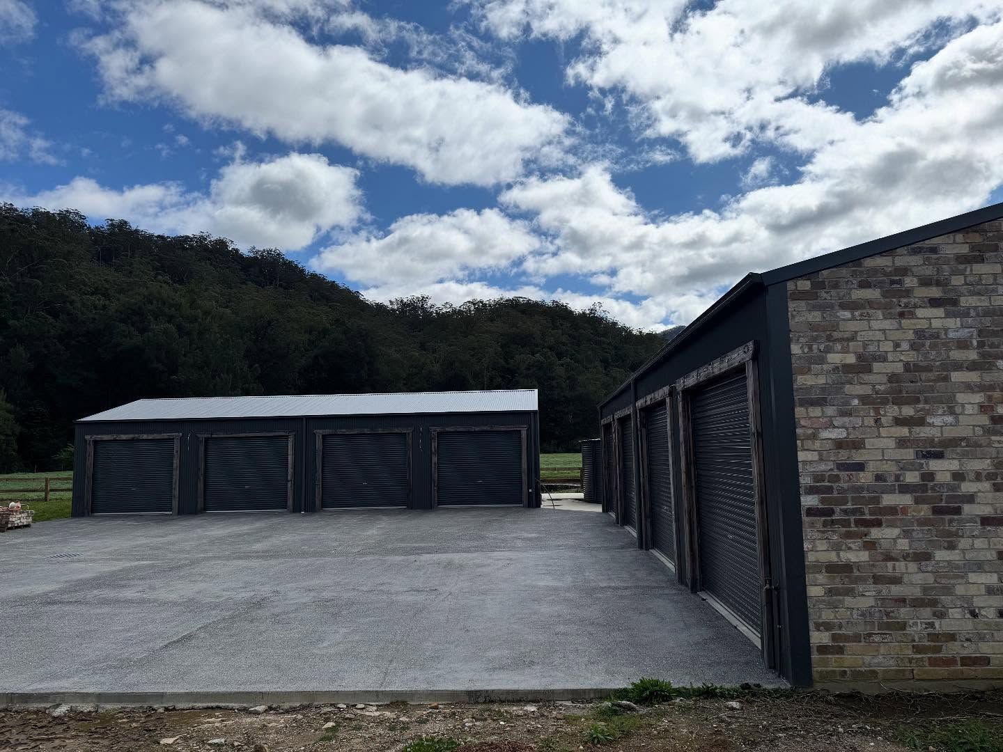 Two dark-coloured garages with roll-up doors, one brick and one wooden, sit on a concrete slab against a wooded hillside.