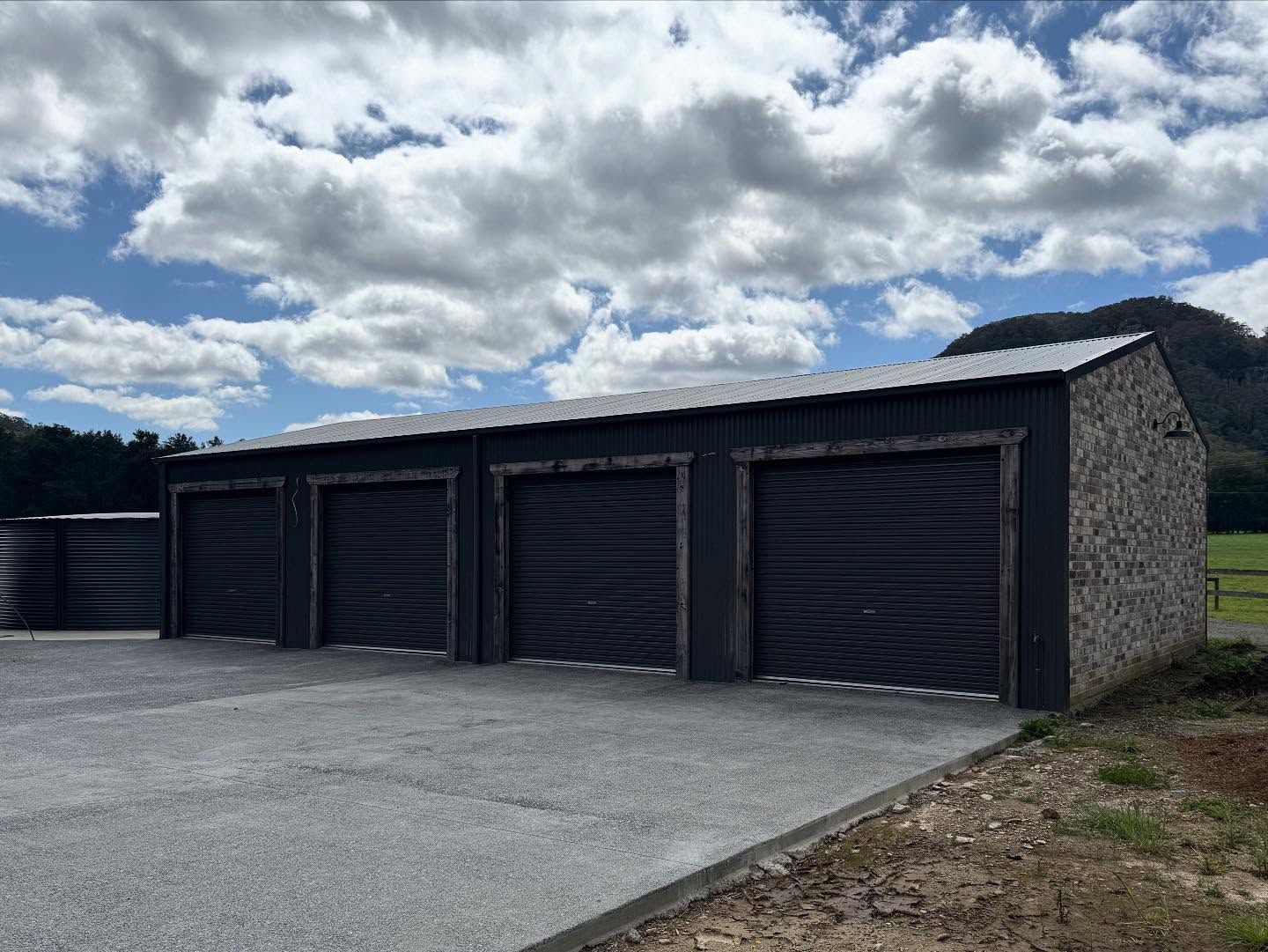 Four-bay garage with black roller doors, gravel driveway, and a cloudy sky background.