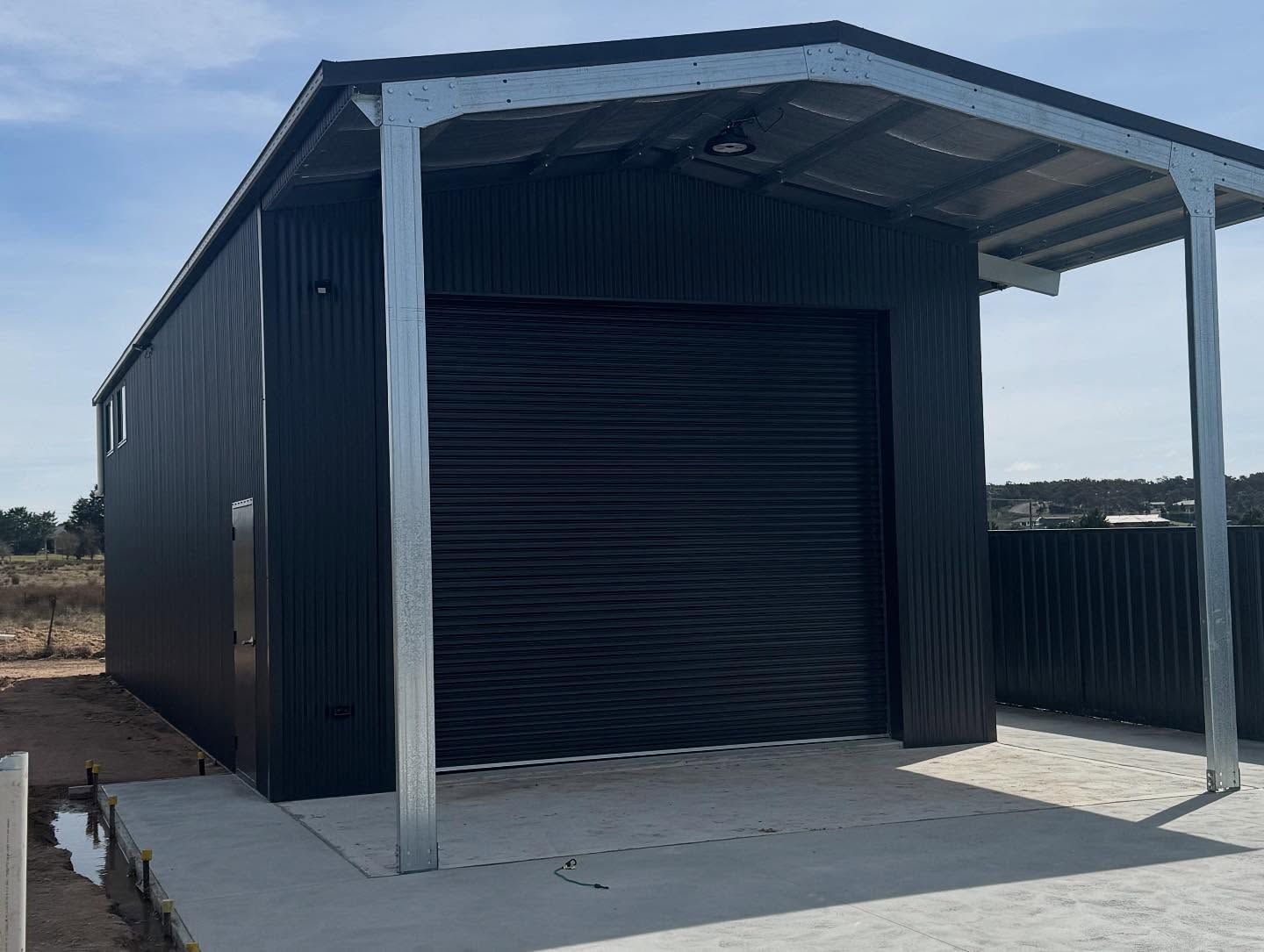 Black metal shed with roller door and attached carport, set on a concrete slab.