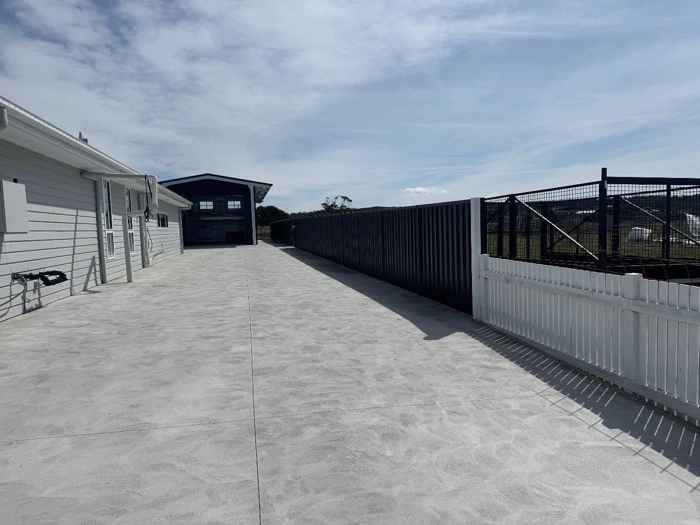 Concrete driveway with a carport and fences on either side under a cloudy sky.