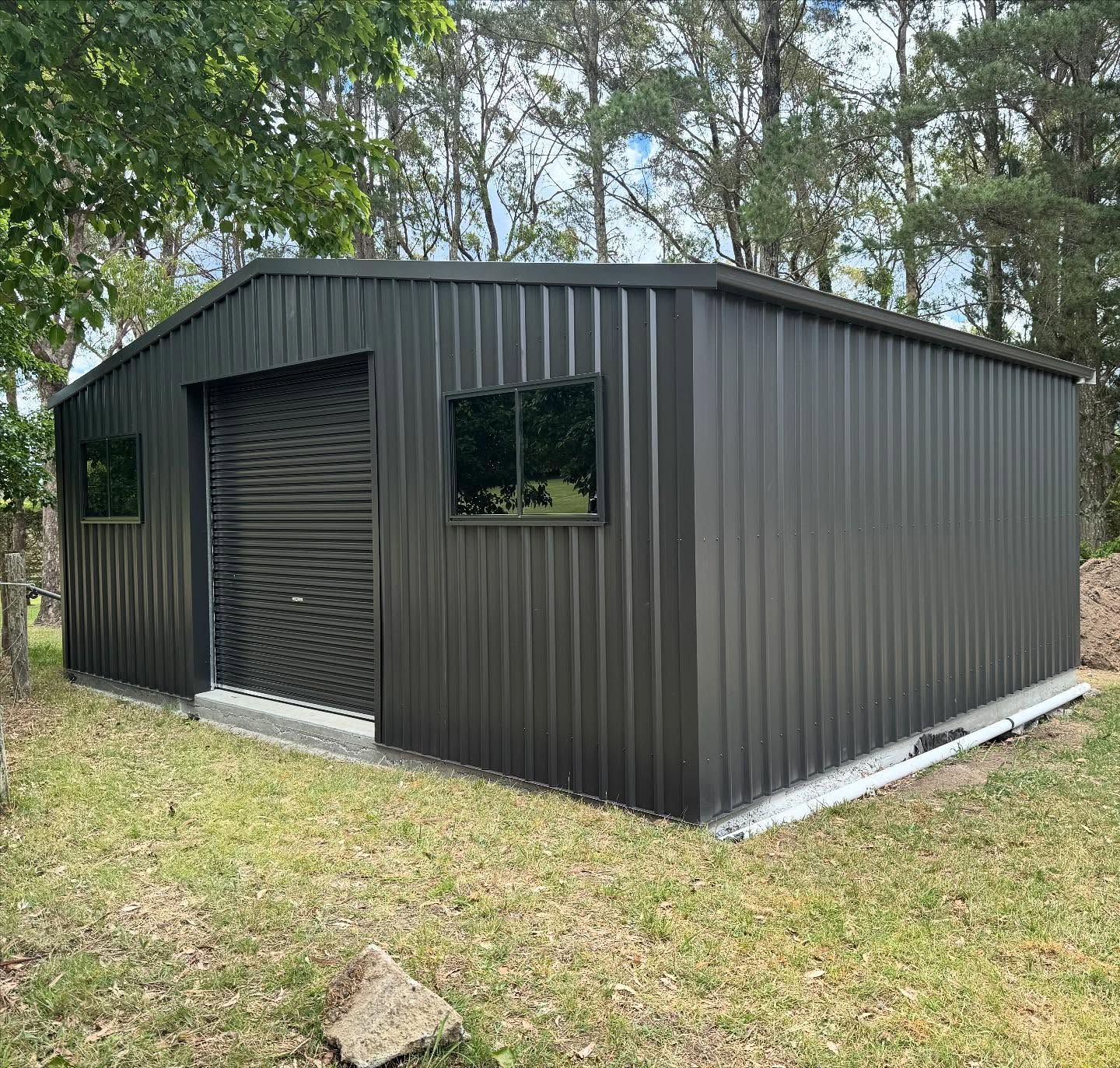 Black metal shed with roller door and windows, set in a grassy area with trees in the background.