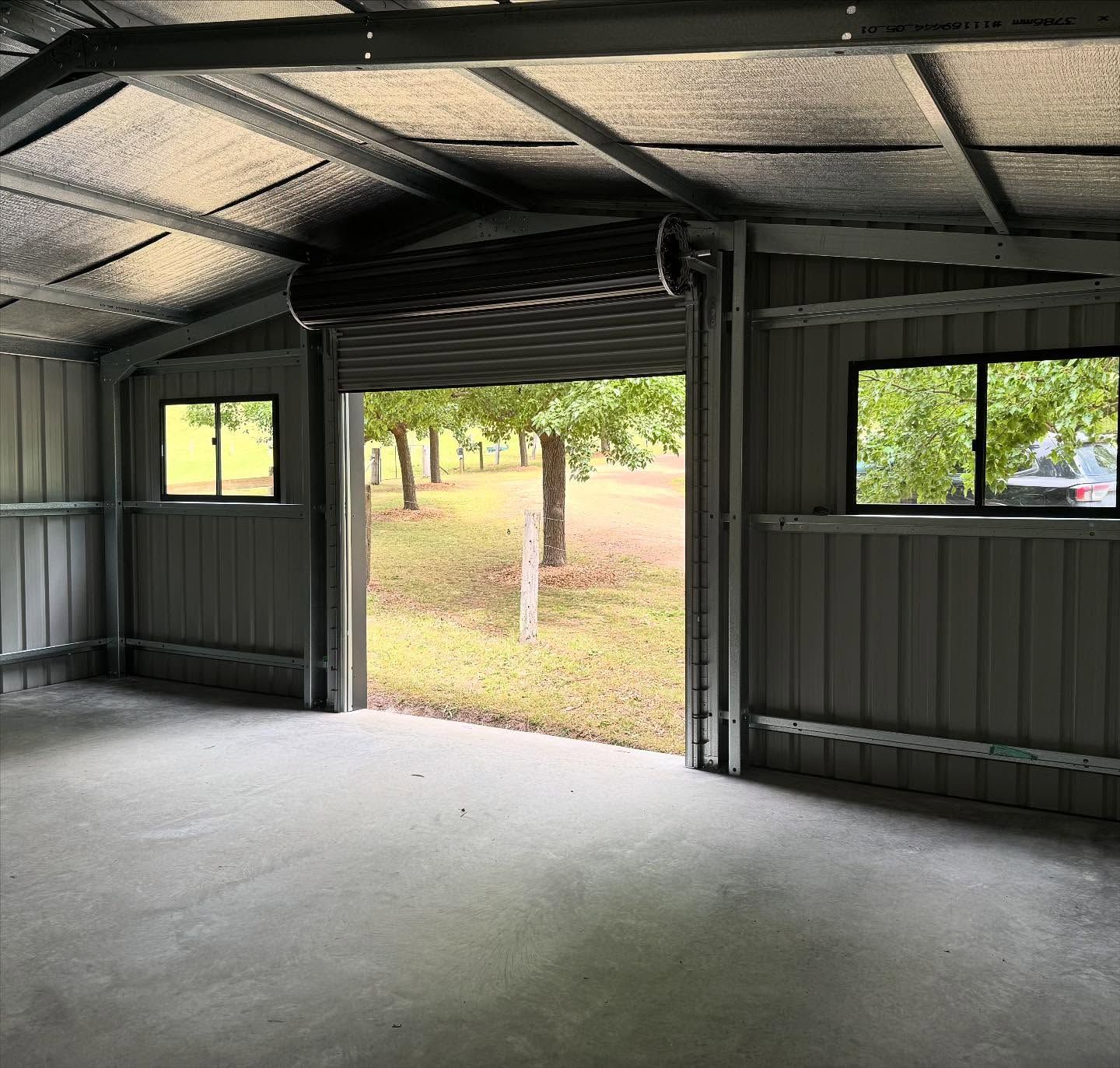 Inside of a metal shed with a roller door open, overlooking a grassy yard and trees.