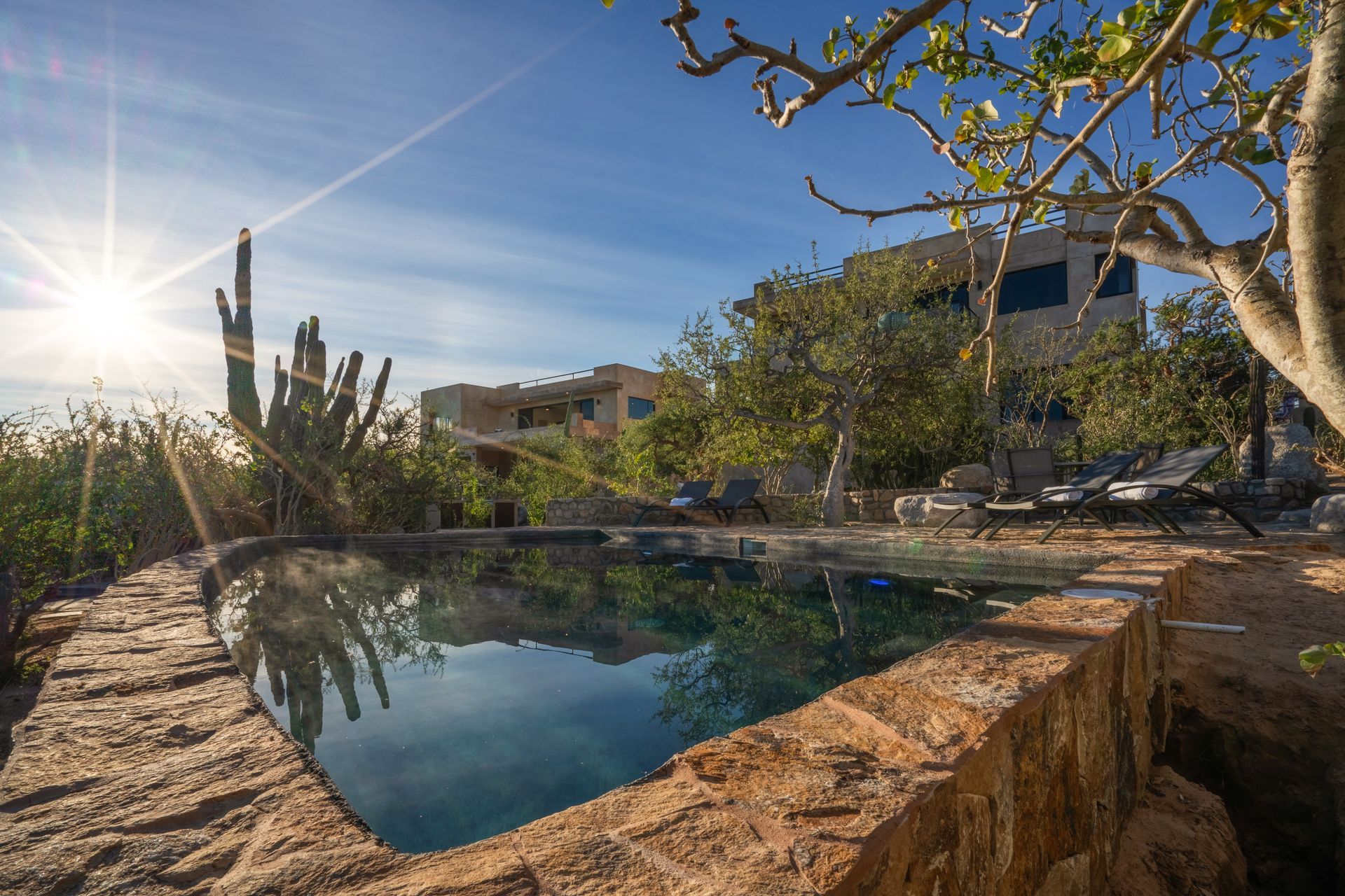Stone-edged pool with sunlit water, overlooking a desert landscape with buildings and cacti under a blue sky.