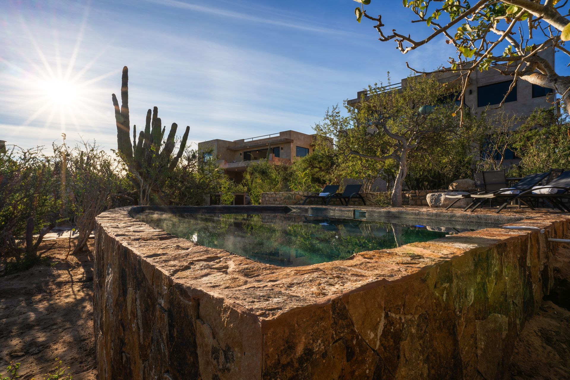 Stone-walled pool with turquoise water, desert landscaping, luxury homes in the background, sunburst sky.