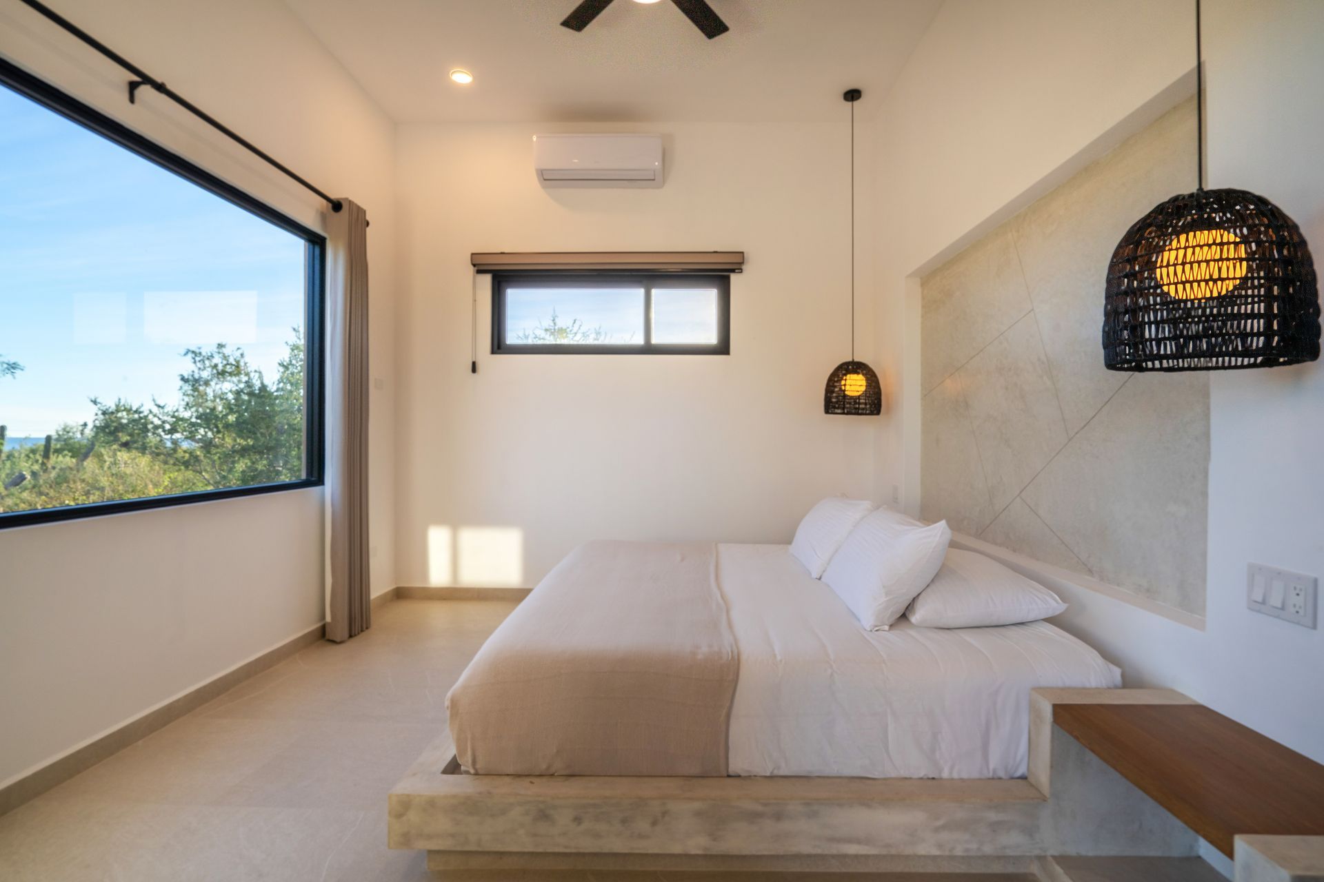 Bedroom with a large window overlooking trees, a bed with white linens, and a hanging light fixture.