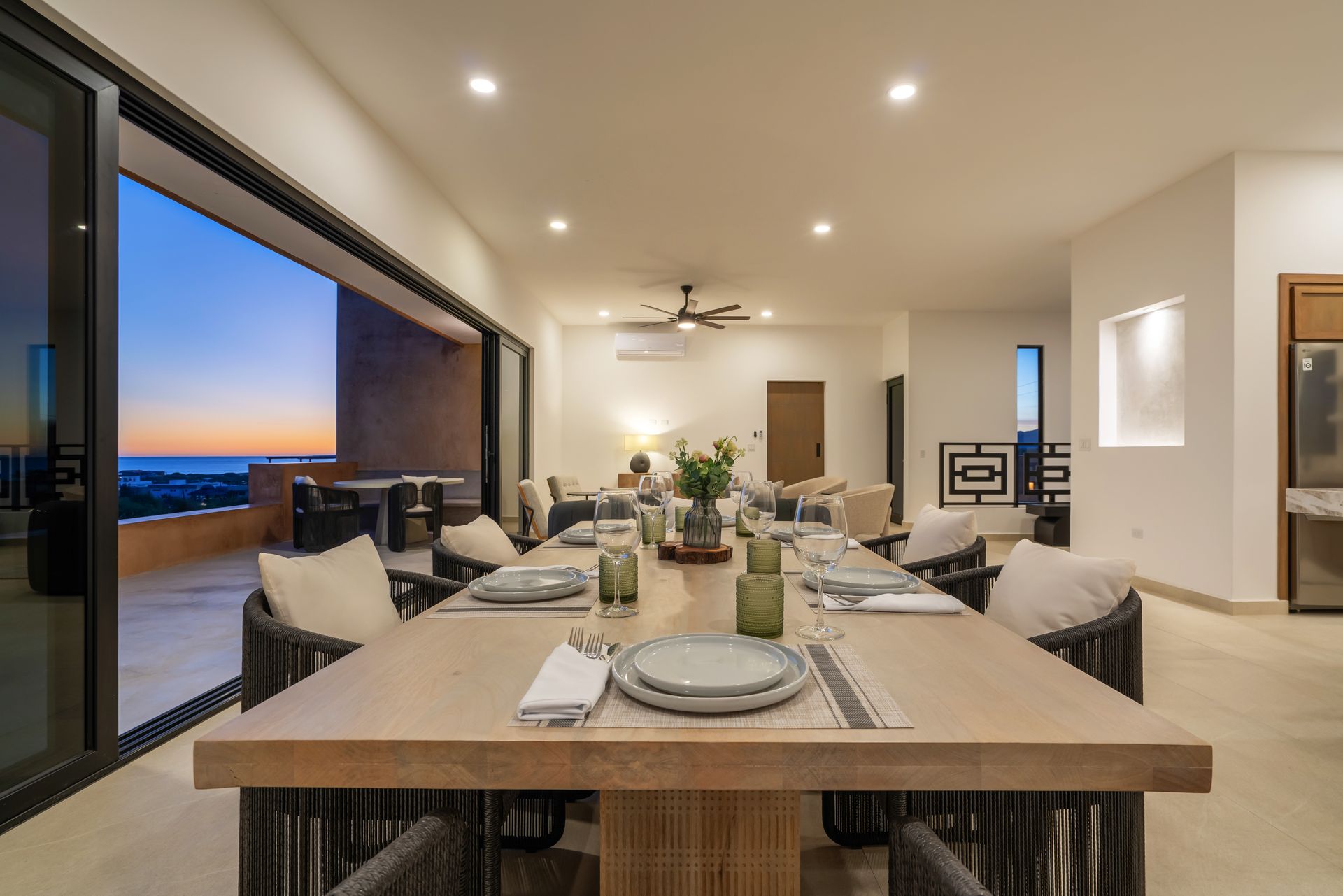 Dining room with long table set for a meal, open to an outdoor patio with ocean view at sunset.