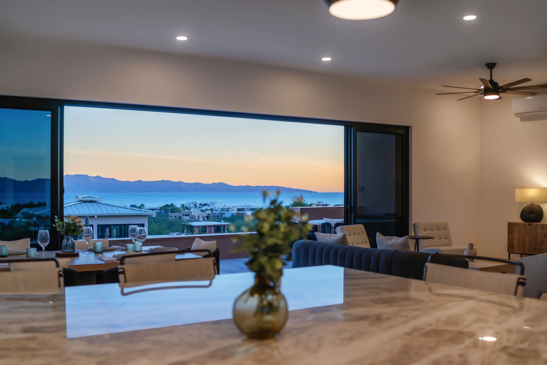 Interior view of a modern home with open sliding doors revealing a scenic sunset over a city and water.