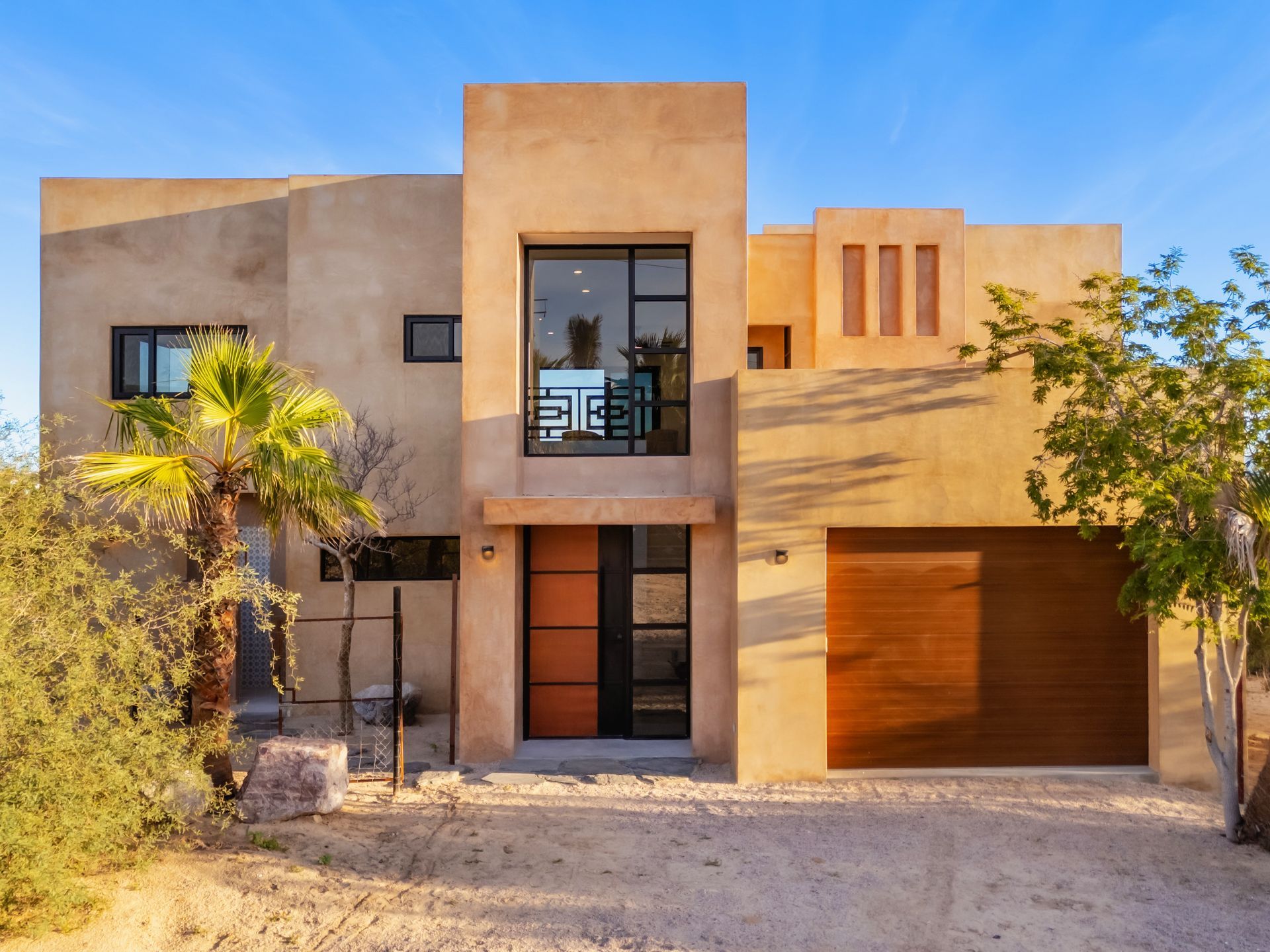 Beige stucco modern house with brown accents, garage, and large front window against blue sky.