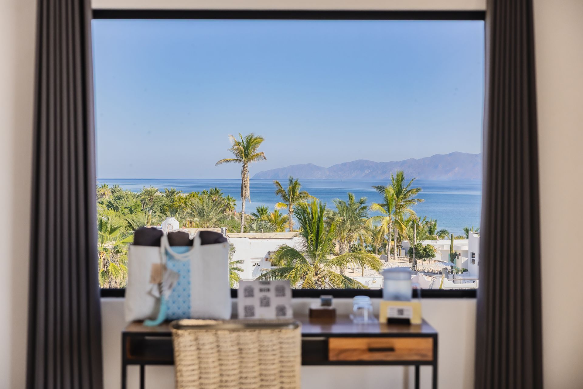 Desk with tote bag, water dispenser, and view of the ocean, palm trees, and mountains framed by a window.