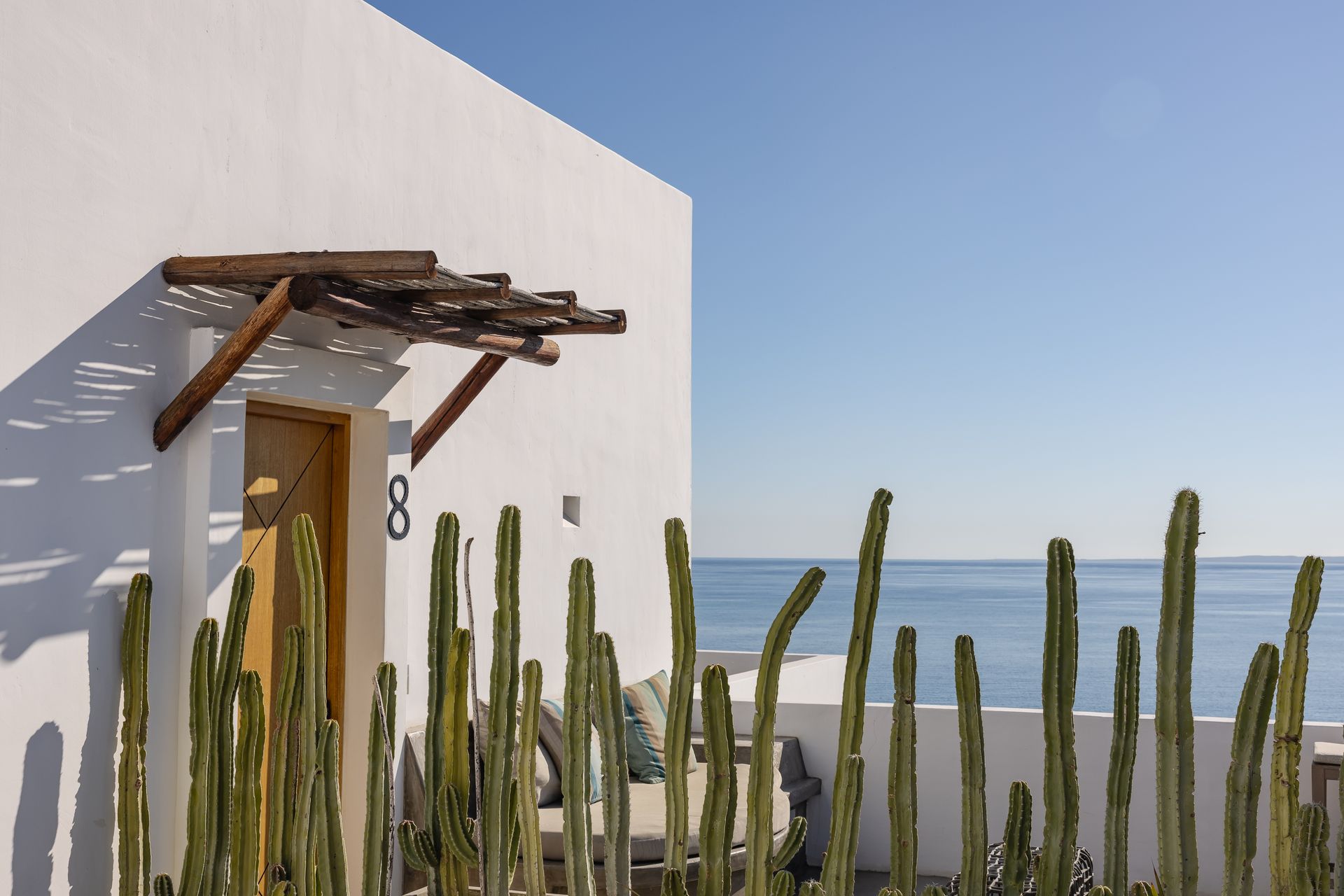 White building with a wooden door under a rustic awning, cactus in front, ocean view.