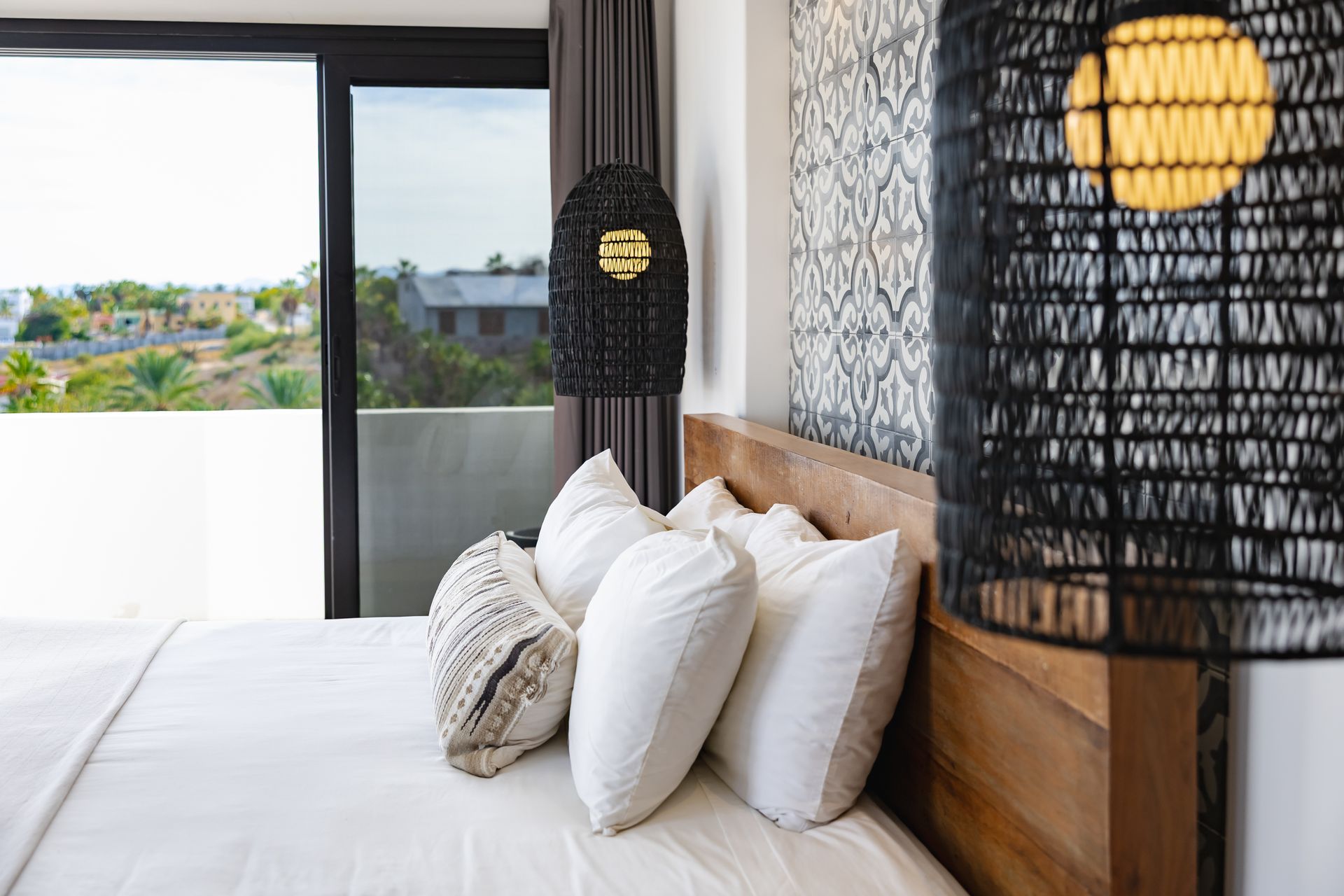 Bedroom with white bedding, pillows, wooden headboard, large window with view, and woven black lamps.