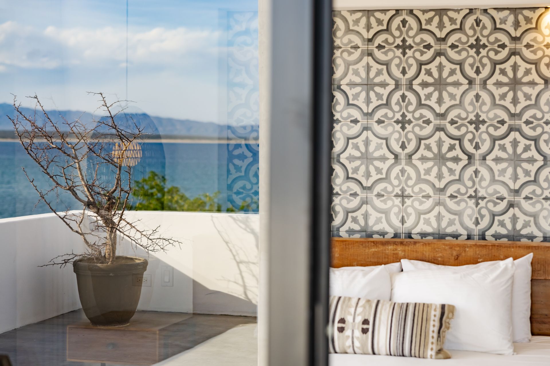 Bedroom with balcony overlooking water; patterned wall, potted plant, white bedding.