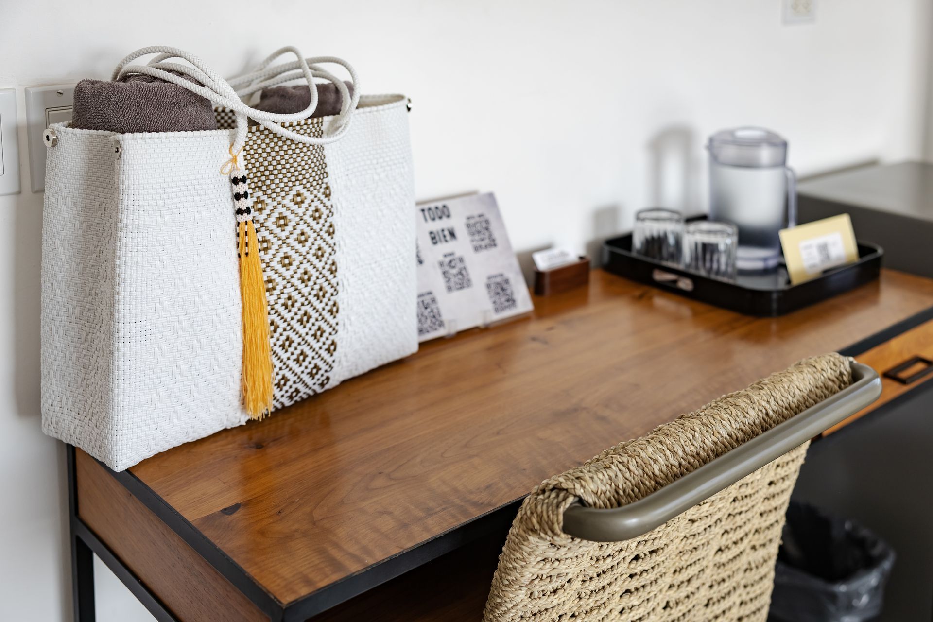 A woven handbag on a desk in a hotel room, towels, and a water carafe are on a tray.