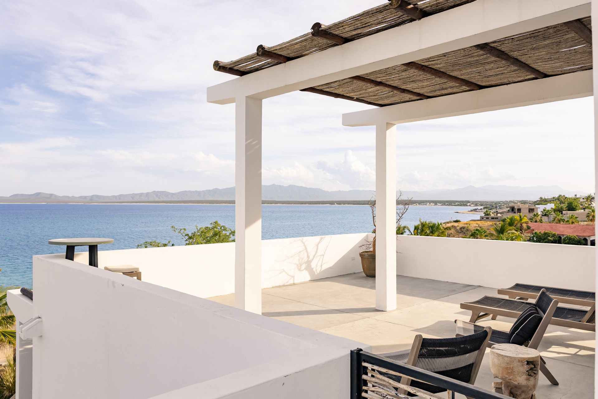 White patio with ocean view, shaded by a wooden pergola; lounge chairs and calm water.