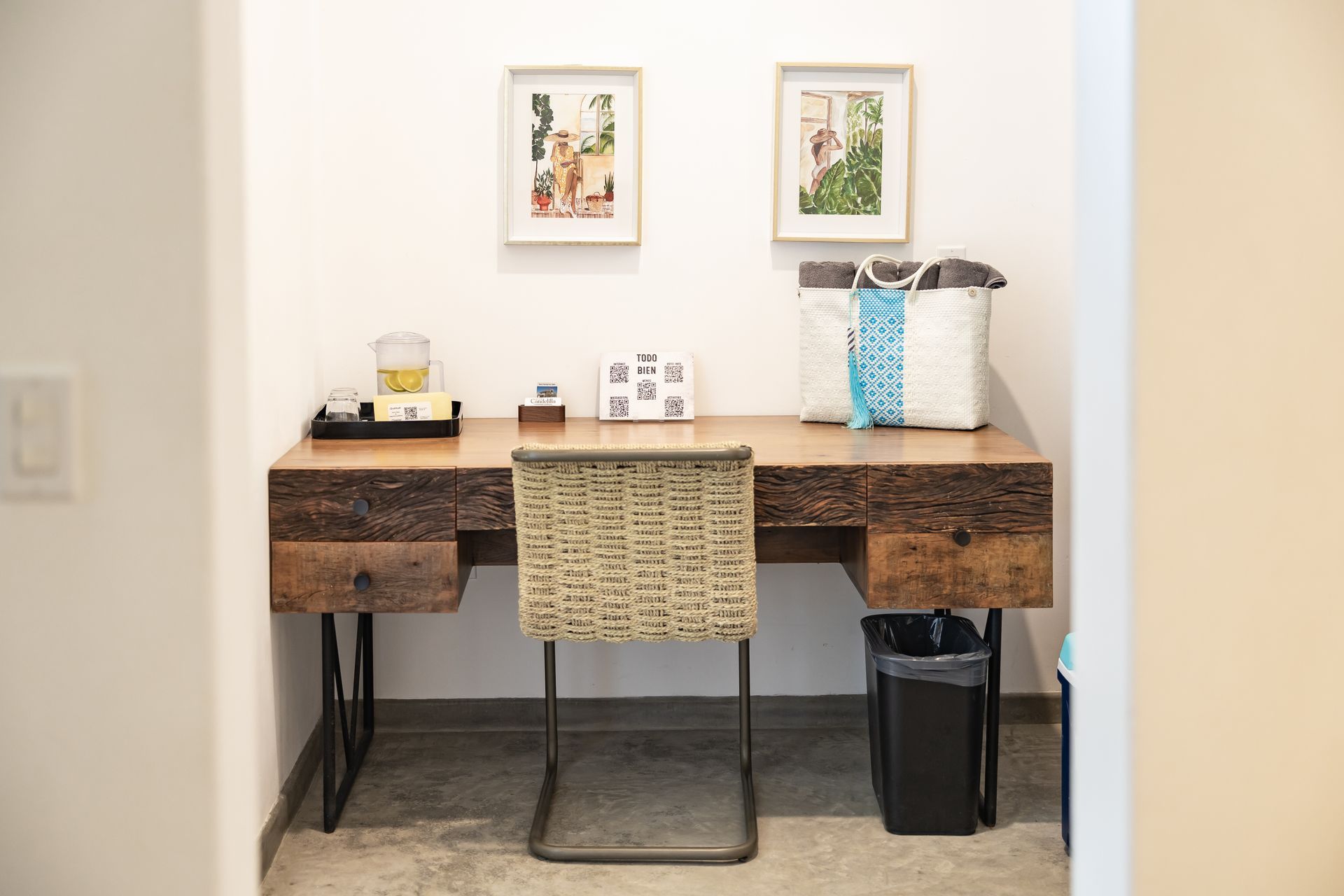 Desk with framed art, chair, tote bag, trash can, and decor in a white-walled alcove.