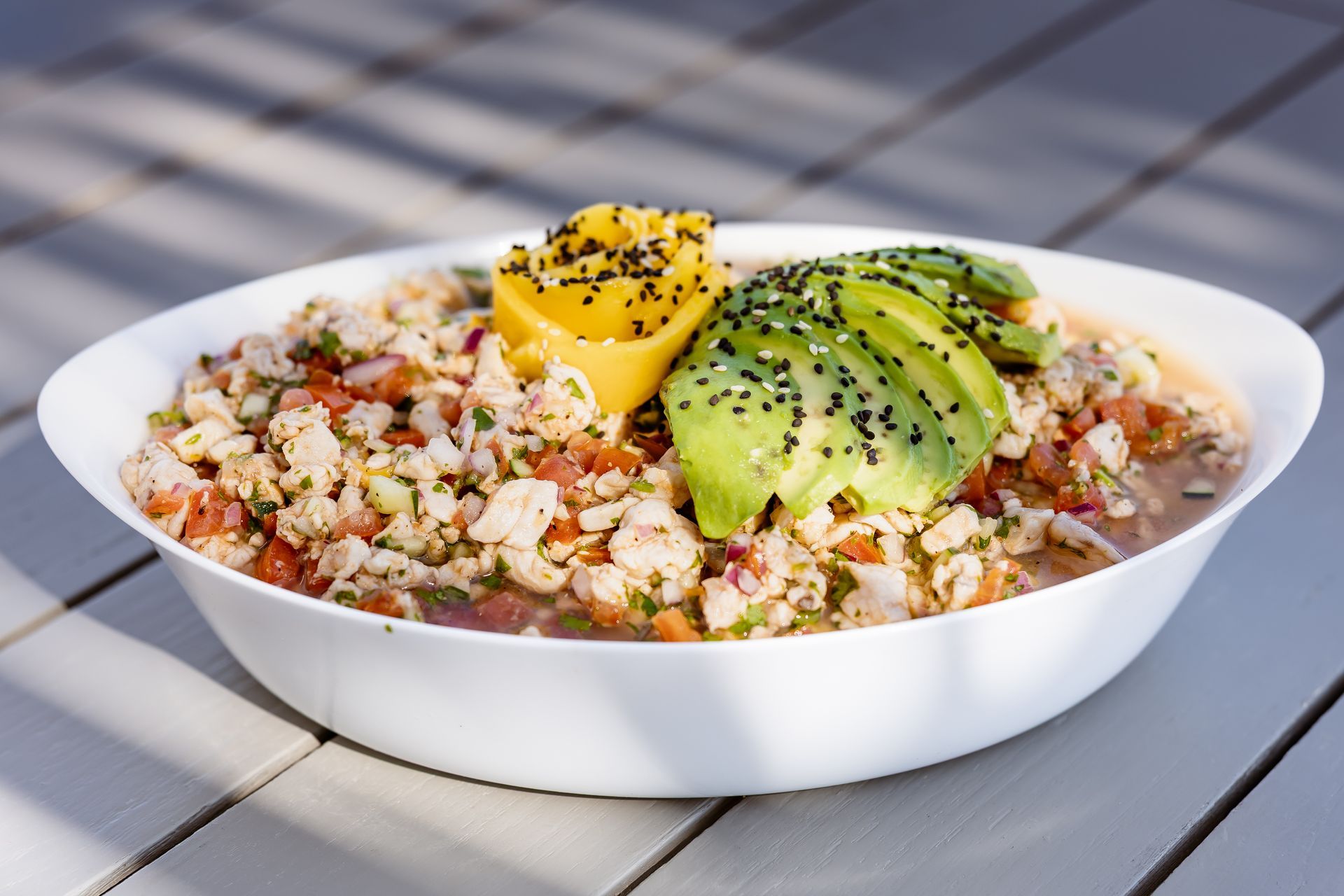 Ceviche in a white bowl, garnished with avocado and mango, sprinkled with black sesame seeds, on a white table.