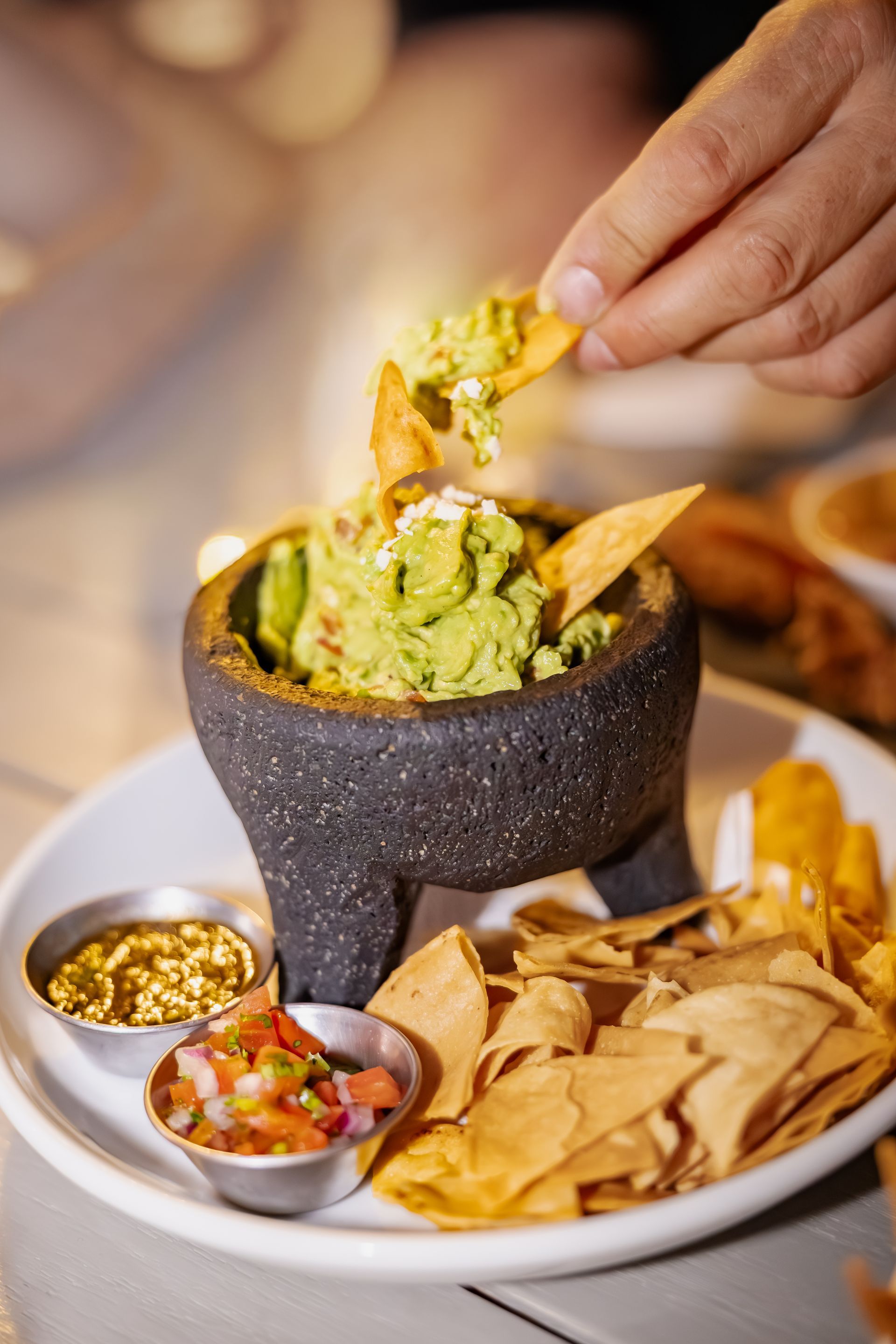 Guacamole in a molcajete, being scooped with a tortilla chip, alongside salsa and chips.