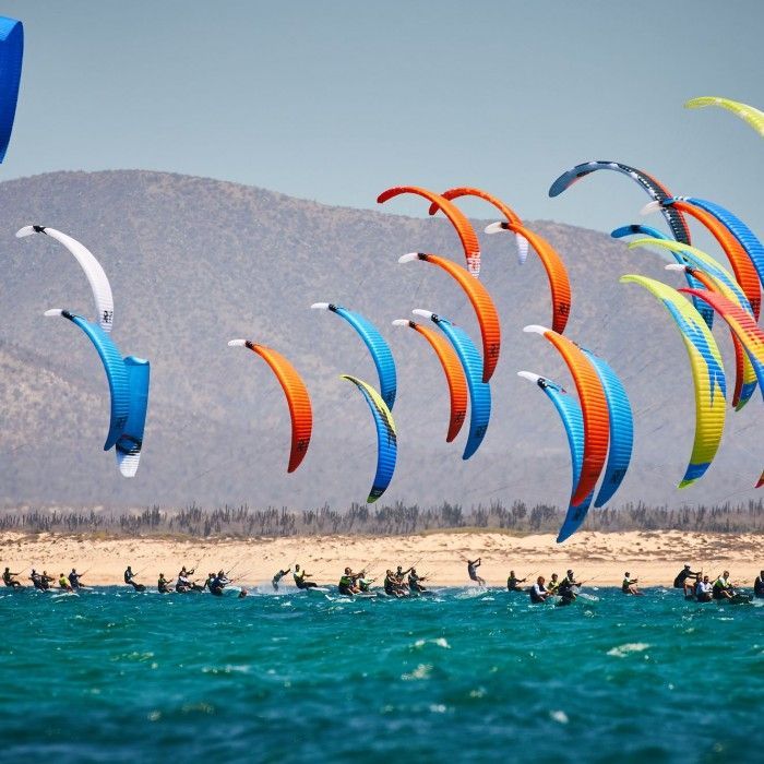Kitesurfers on a beach with colorful kites in the sky, mountains in the background.