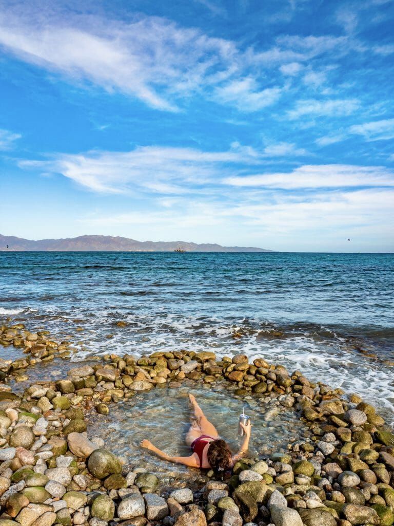 Woman relaxes in a stone-lined pool on a beach, turquoise water, blue sky with clouds, mountains in the distance.