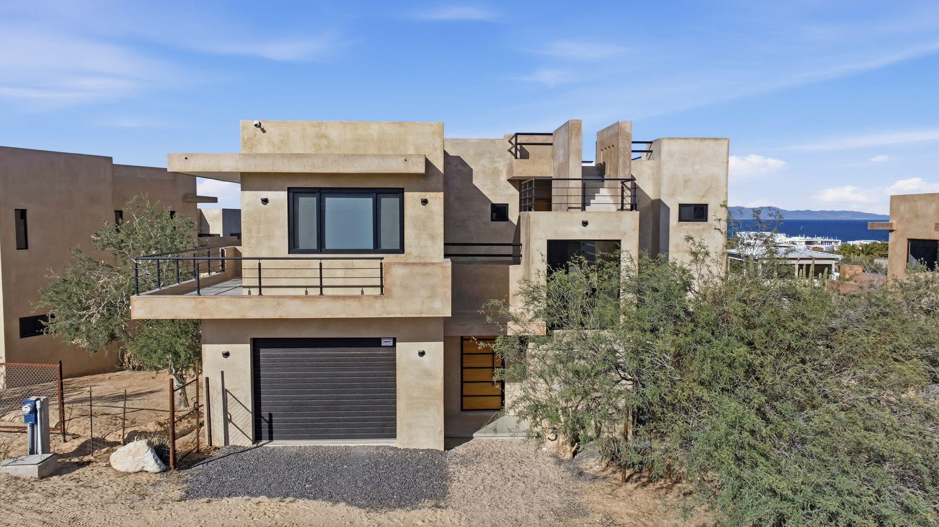 Modern two-story beige house with a dark garage door, balcony, and nearby shrubs, set against a blue sky.