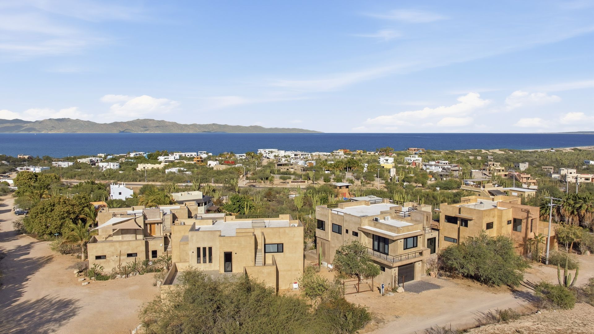 A town with beige buildings and green trees overlooks a body of water under a blue sky.