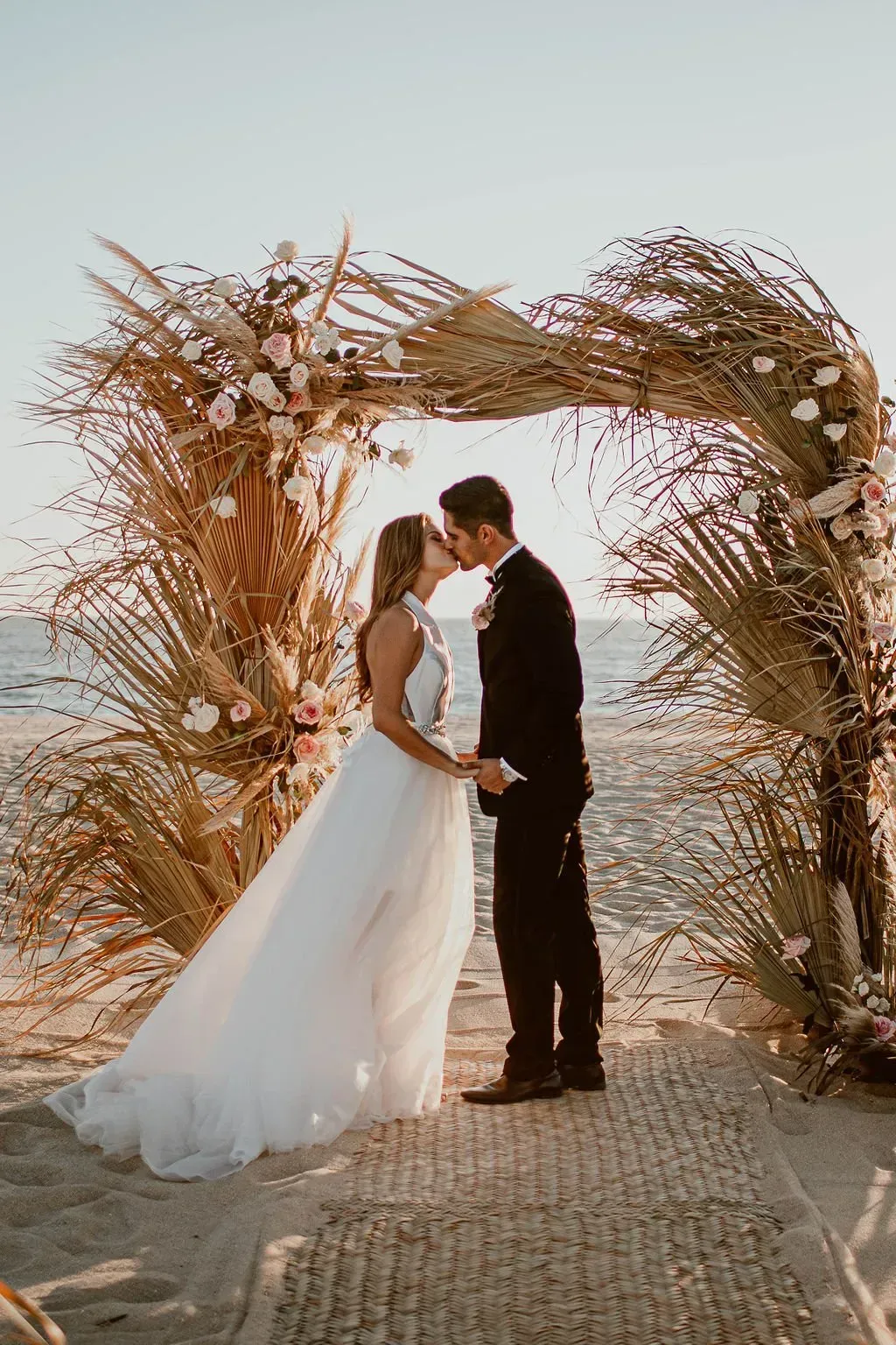 Couple kissing under floral wedding arch on a beach. Sunny day. The bride wears a white dress. Groom in a black suit.