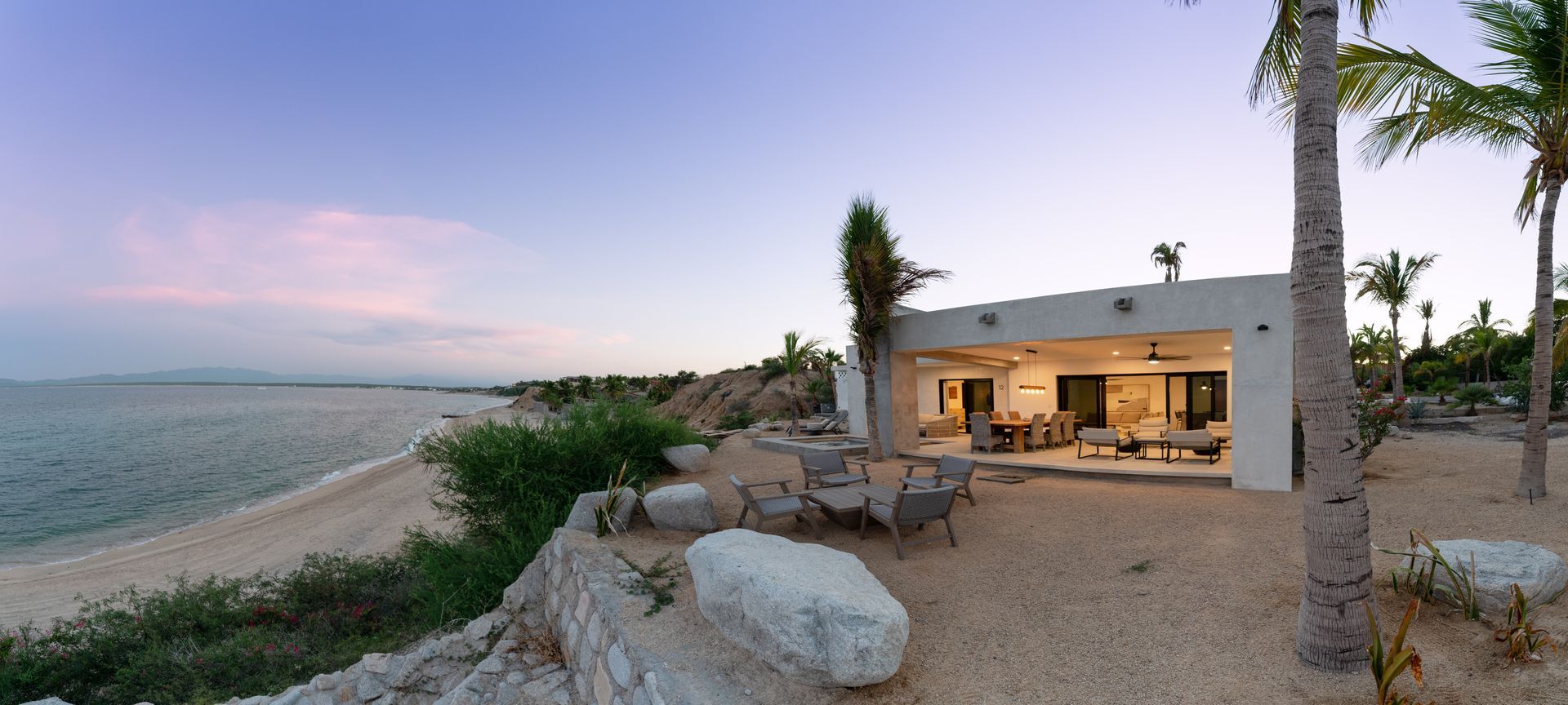 Beachfront house at sunset. Sandy shore, blue water, white building, palm trees, outdoor seating area.