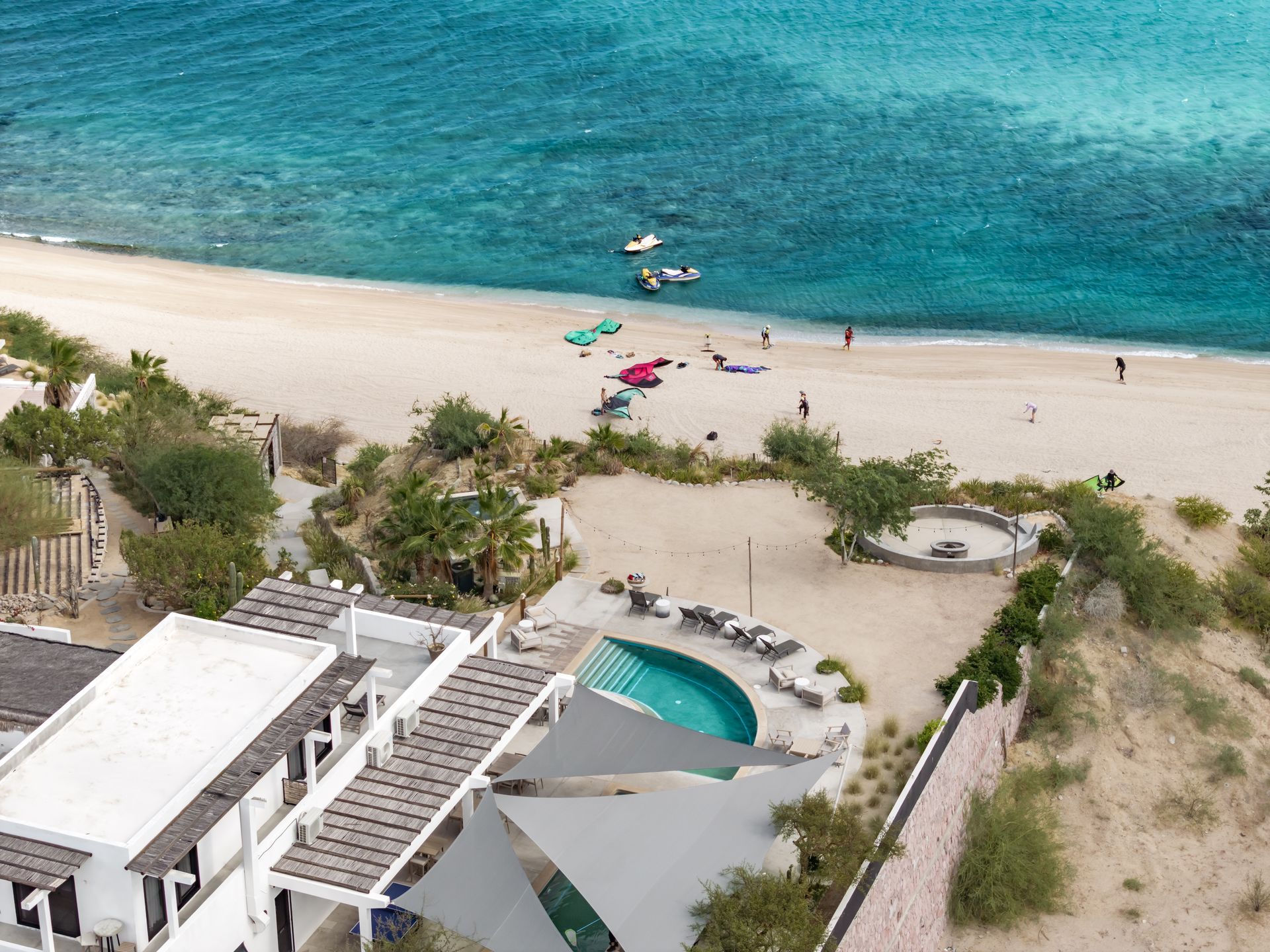 Beachfront resort with white buildings, blue pool, and sandy beach with people and watercraft.
