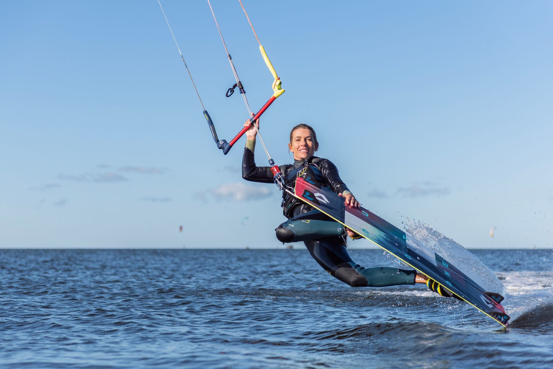 Person kitesurfing on water, holding kite bar, smiling, bright blue sky.