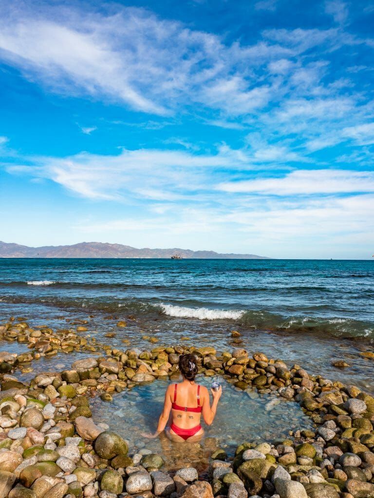 Woman in a red bikini sits in a small, rock-lined pool on a beach. Ocean and mountains in the background. Sunny day.
